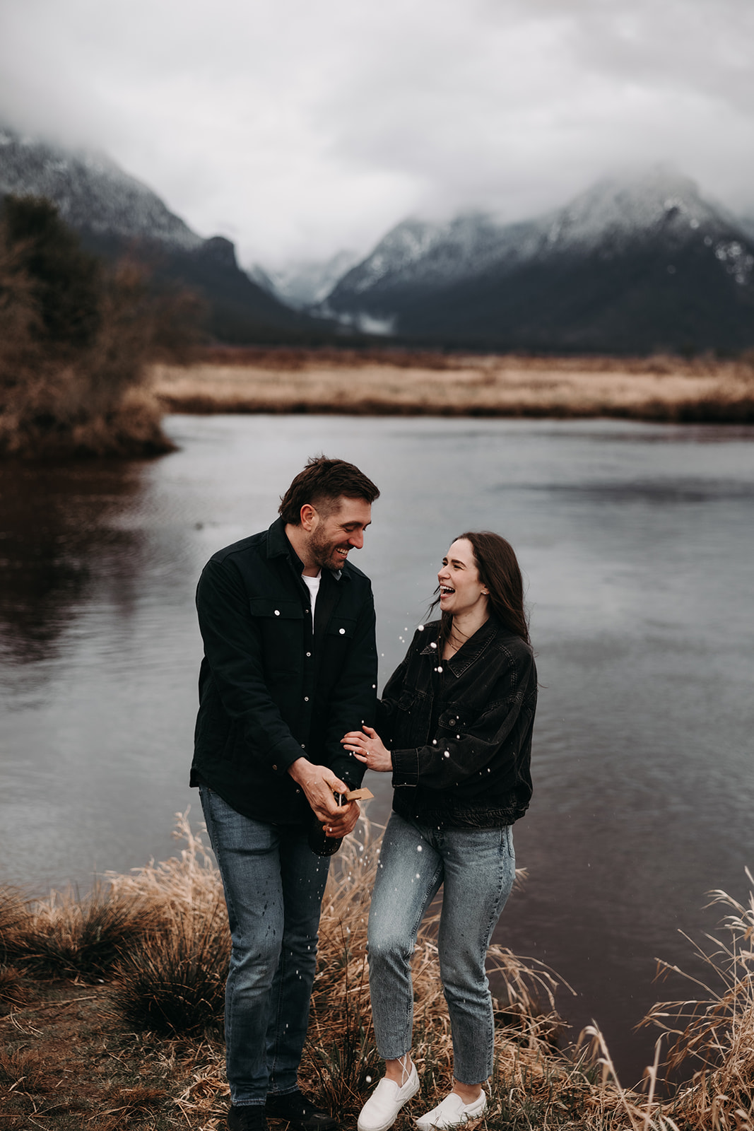  In this sweet fall engagement photo, the couple shares a tender moment by the river, smiling and holding hands as they gaze into each other's eyes. The lush, muted colors of the autumn landscape complement their relaxed, casual outfits, showcasing the beauty of love in the great outdoors.