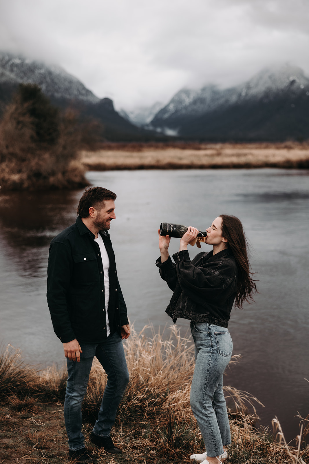 The woman playfully drinks from a bottle while her partner watches with a smile, creating a fun and candid moment in their fall engagement photos. The background features a serene river and snow-capped mountains, highlighting the picturesque setting of Pitt Lake and the vibrant connection they share.