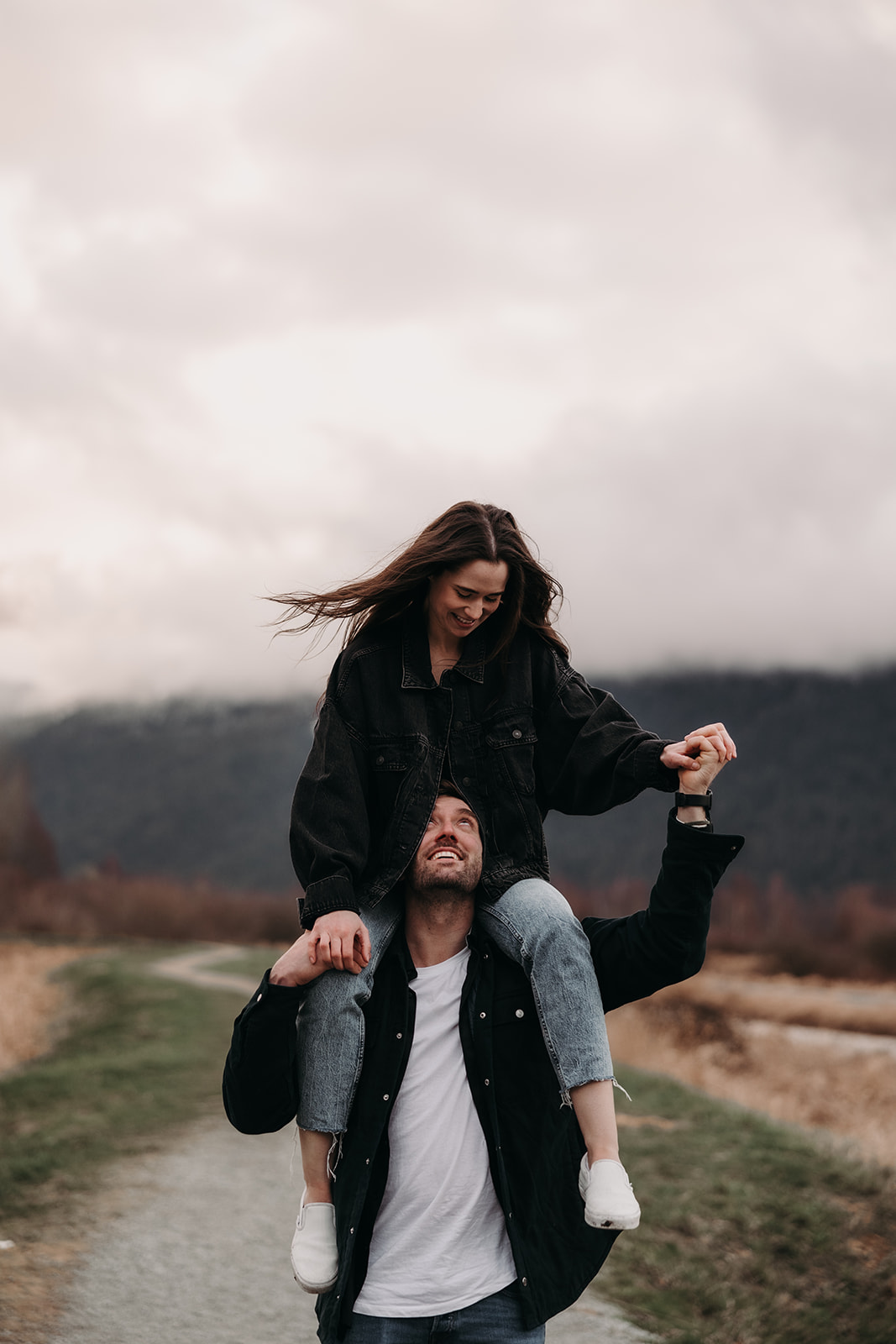  In a playful scene, the woman sits on her partner's shoulders, laughing joyfully as he holds her hands, showing off her engagement ring. They are surrounded by beautiful fall colors and mountains in the distance, adding a romantic touch to their fall engagement photos. This image radiates fun and excitement, perfectly capturing the couple's adventurous spirit and love for each other.