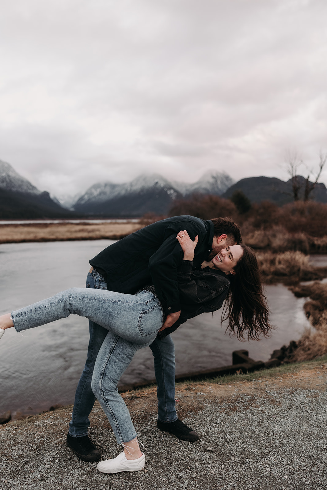 A couple joyfully poses near a tranquil river, with the woman being playfully lifted by her partner. She smiles brightly, her hair flowing with the movement, while he has a cheeky grin on his face. The scenic backdrop features misty mountains and soft clouds, creating an ideal setting for their fun fall engagement photos that capture the spirit of adventure and love.