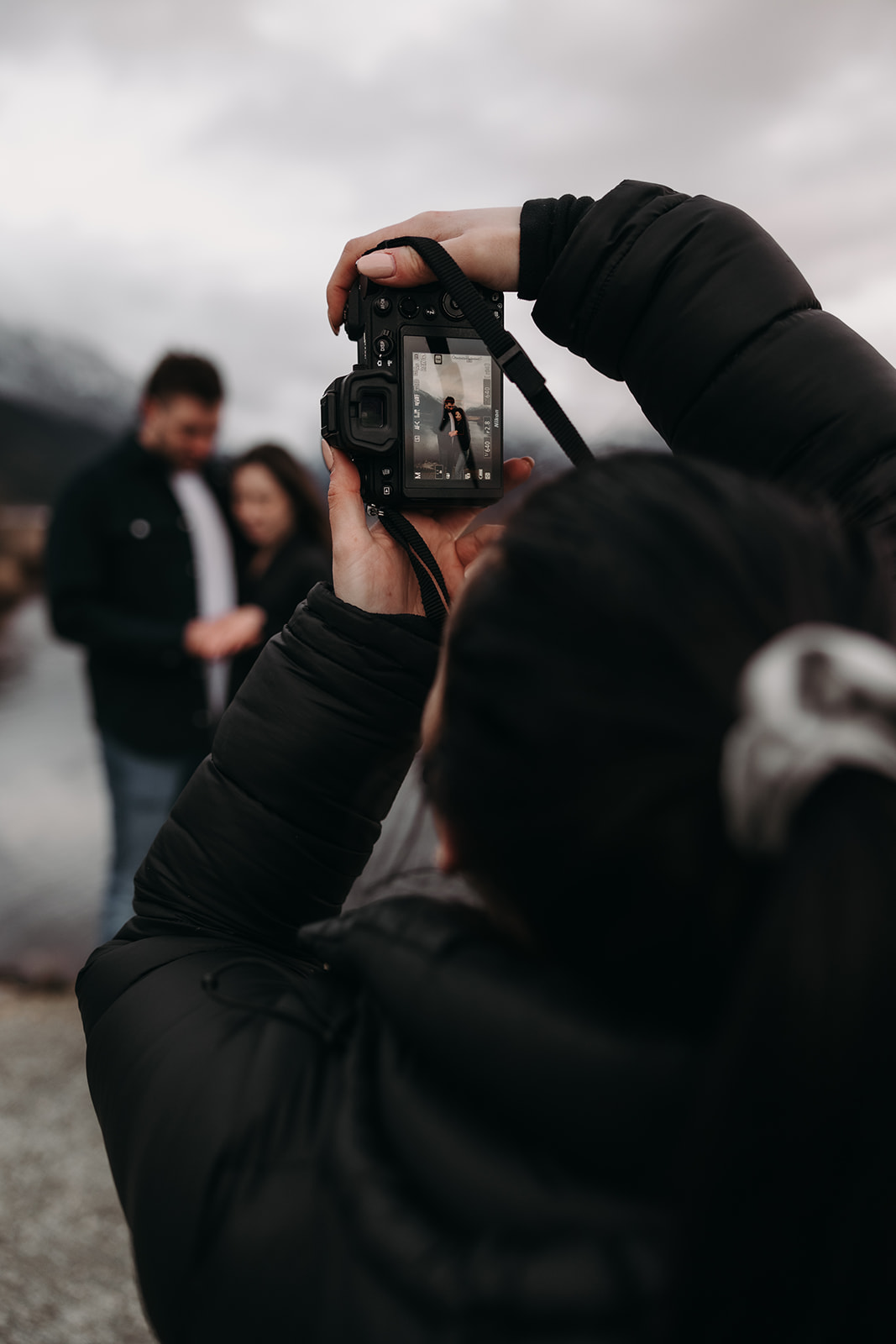 A candid shot of the couple in the foreground with the photographer's hand holding a camera in the background. They share a sweet moment, surrounded by the stunning landscape of Pitt Lake, showcasing the joyful spirit of their fall engagement photos 