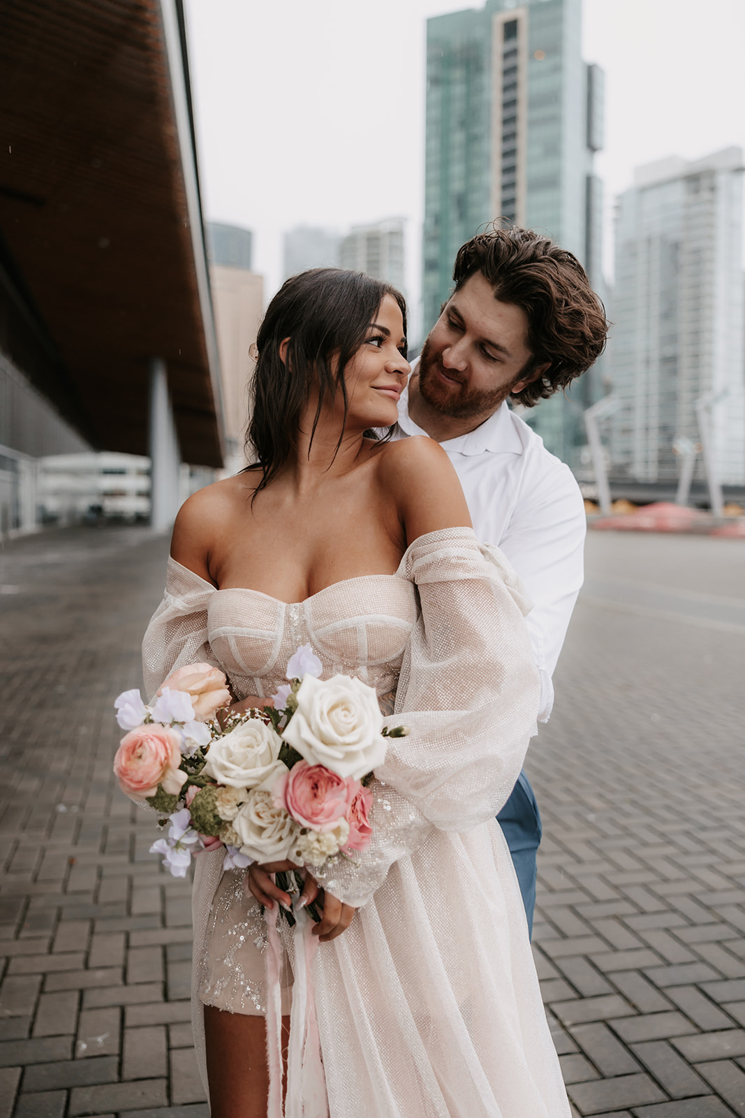 Couple in downtown Vancouver sharing a romantic moment under the architecture of the city, with the bride holding her bouquet of pink roses.