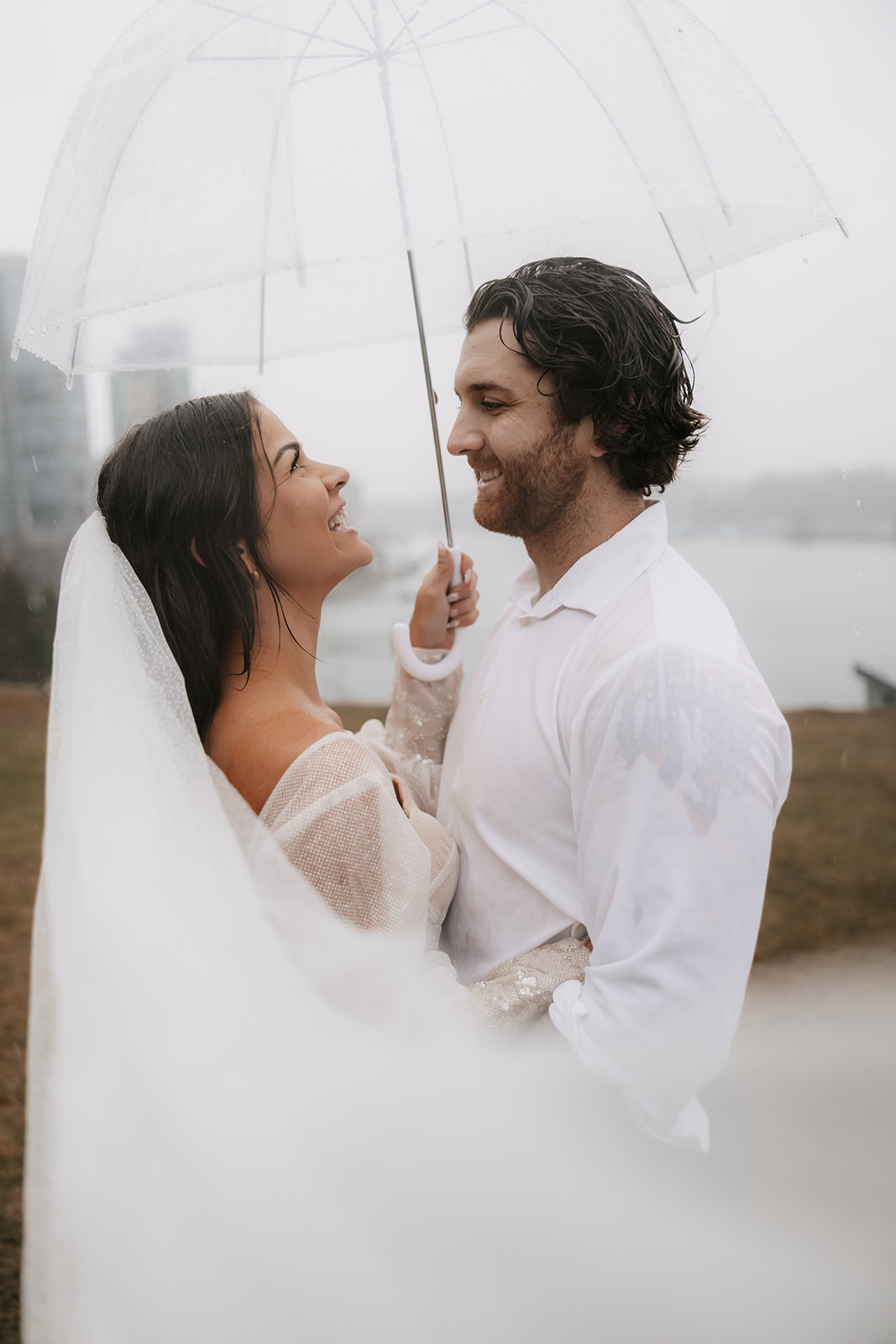 Bride and groom laughing together under an umbrella on a rainy day in Vancouver, with the Vancouver skyline as a backdrop during their elopement session.