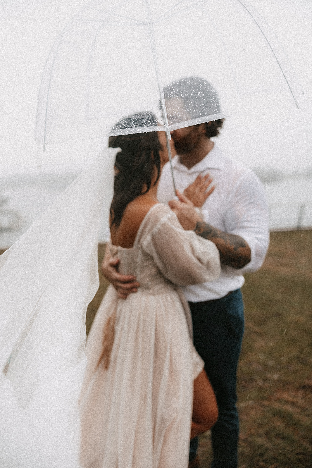 Couple sharing a kiss under a transparent umbrella during their Vancouver Elopement, with rain falling around them and the cityscape in the distance.