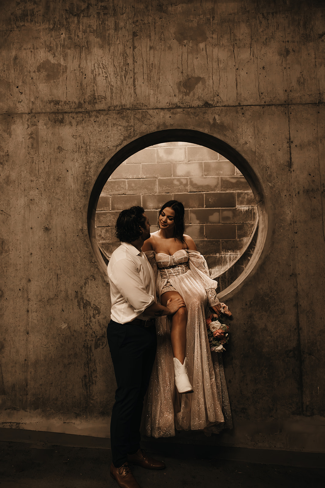 Bride sitting on a cement wall and she glances at her groom in an underground parking garage