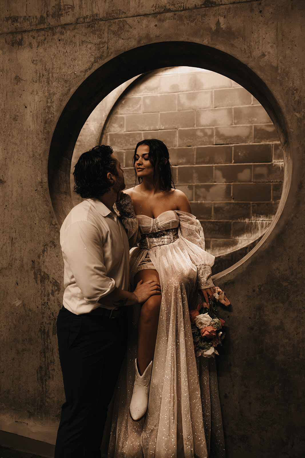 Bride sitting on a cement wall and she glances at her groom in an underground parking garage