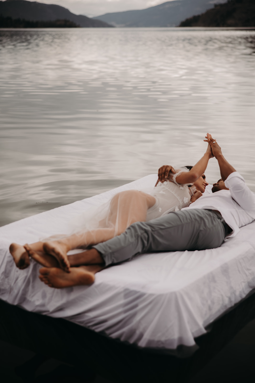 A playful shot of the couple lying on a bed floating on Kalamalka Lake, captured during their Okanagan elopement, surrounded by mountains and misty skies.