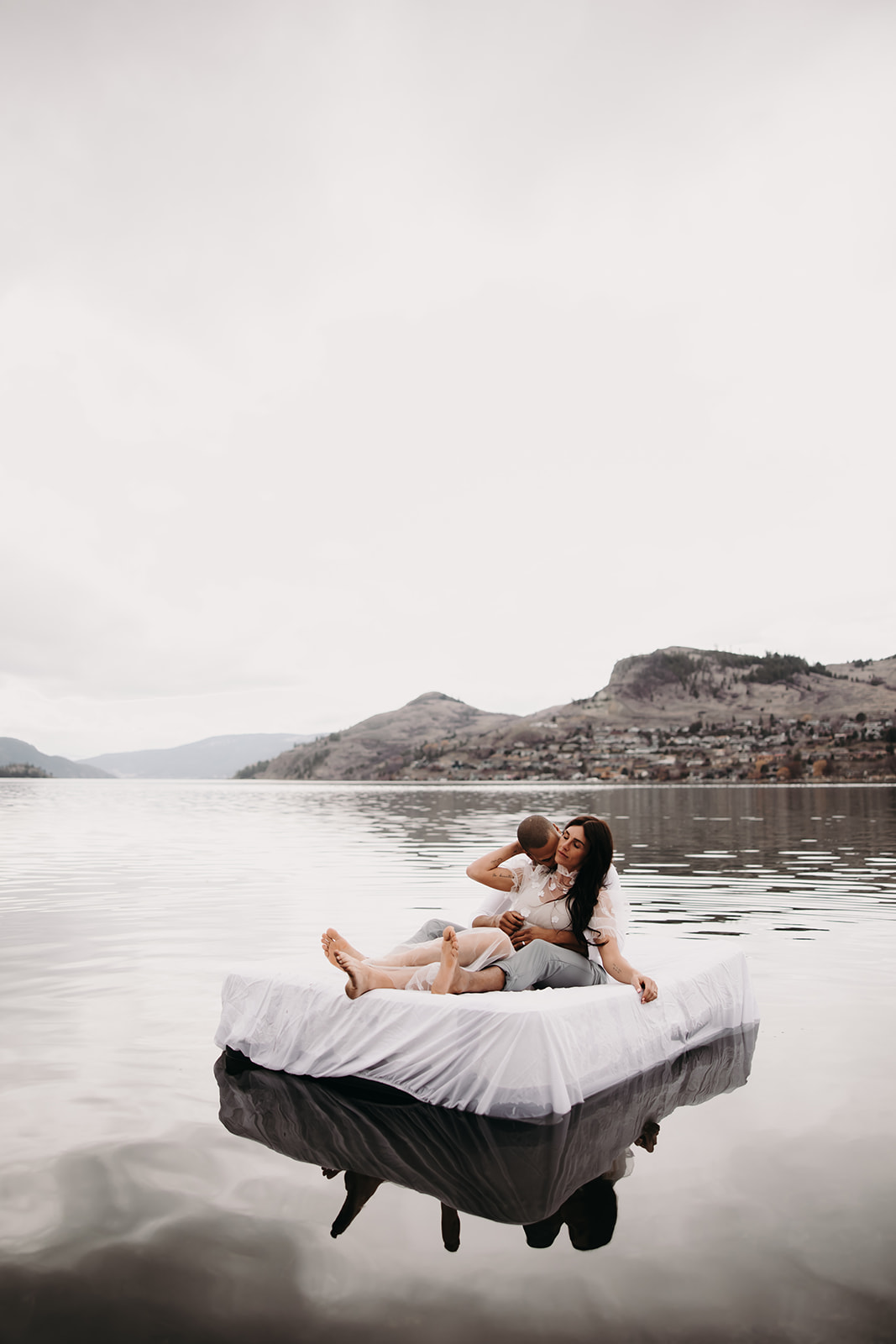 A playful shot of the couple lying on a bed floating on Kalamalka Lake, captured during their Okanagan elopement, surrounded by mountains and misty skies.