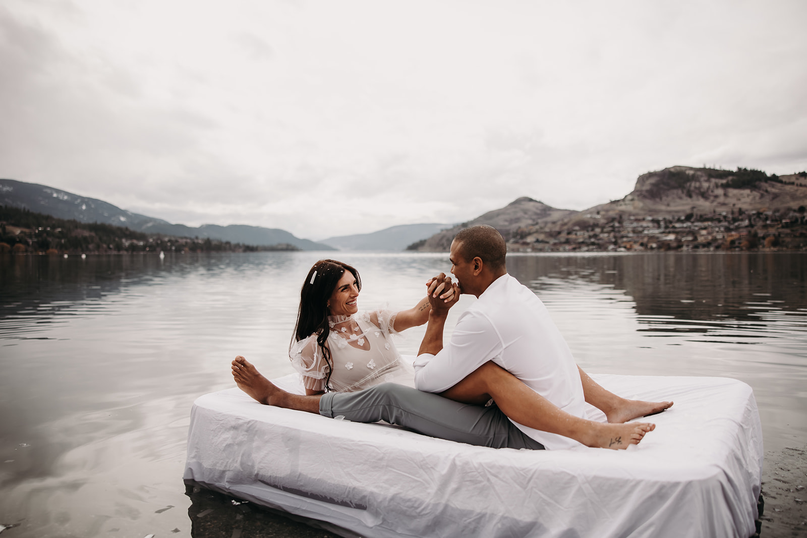 A couple relaxes on a floating bed in Kalamalka Lake, sharing a sweet moment during their adventurous Okanagan elopement with a backdrop of rolling hills.