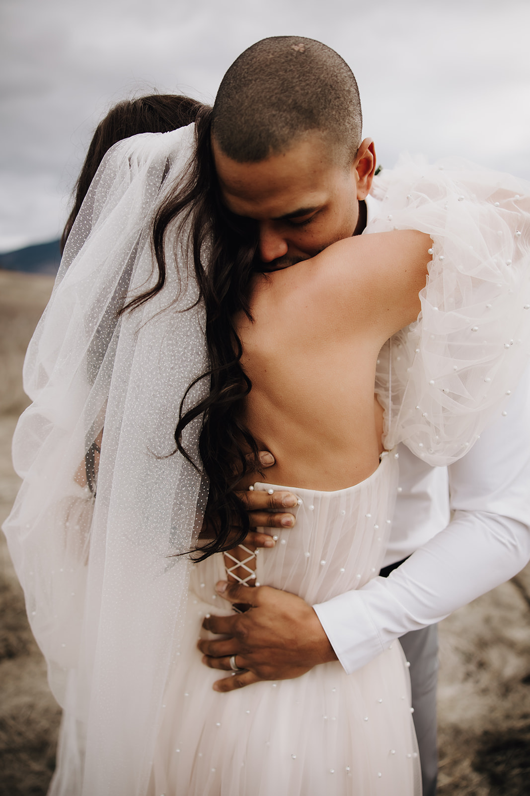 The couple shares a heartfelt hug, with the bride’s veil flowing in the wind and the mountain backdrop of their Okanagan elopement enhancing the intimate moment.