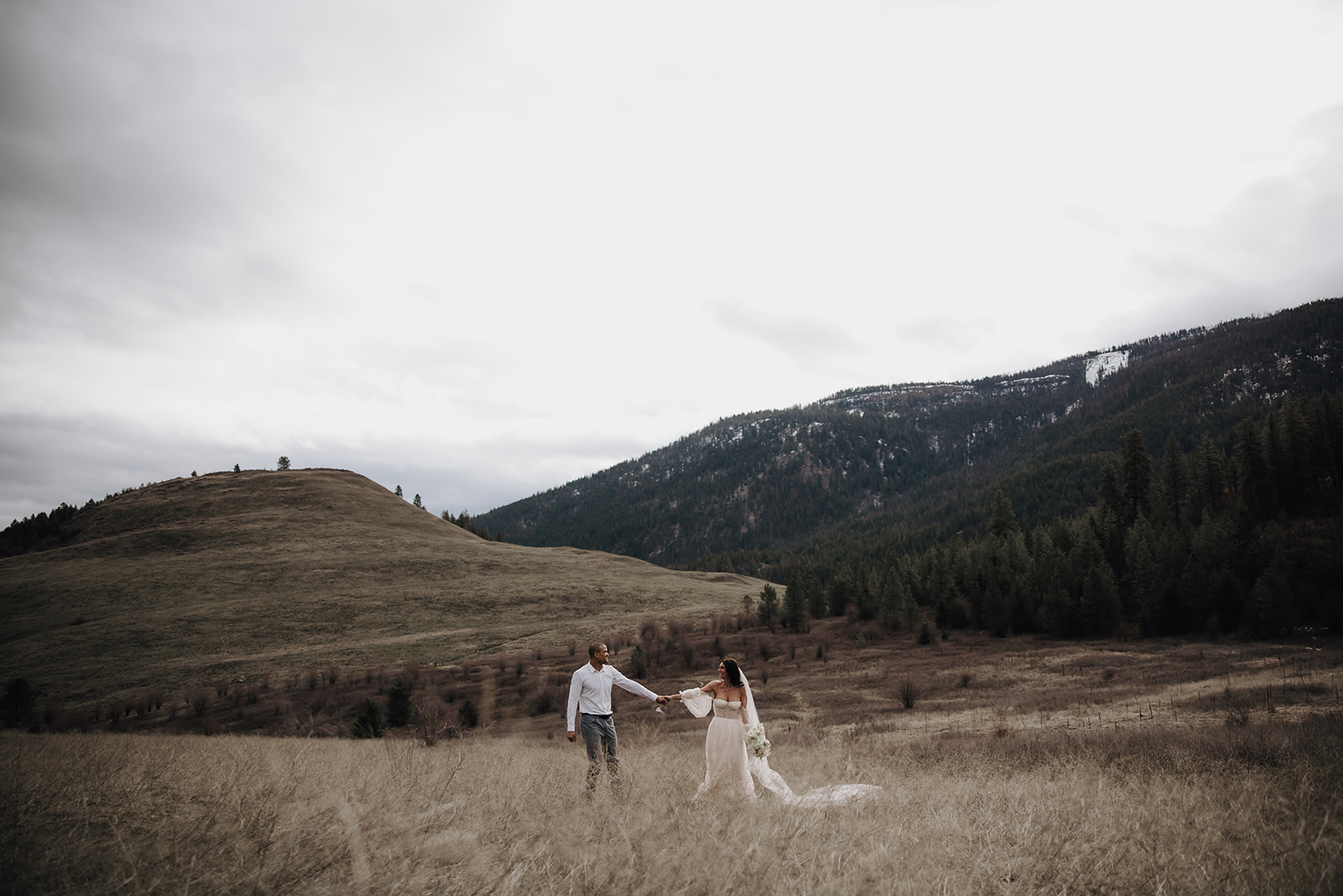 A couple walks hand-in-hand through the fields of Okanagan, surrounded by lush hills and mountains, celebrating their intimate elopement in the stunning landscape.