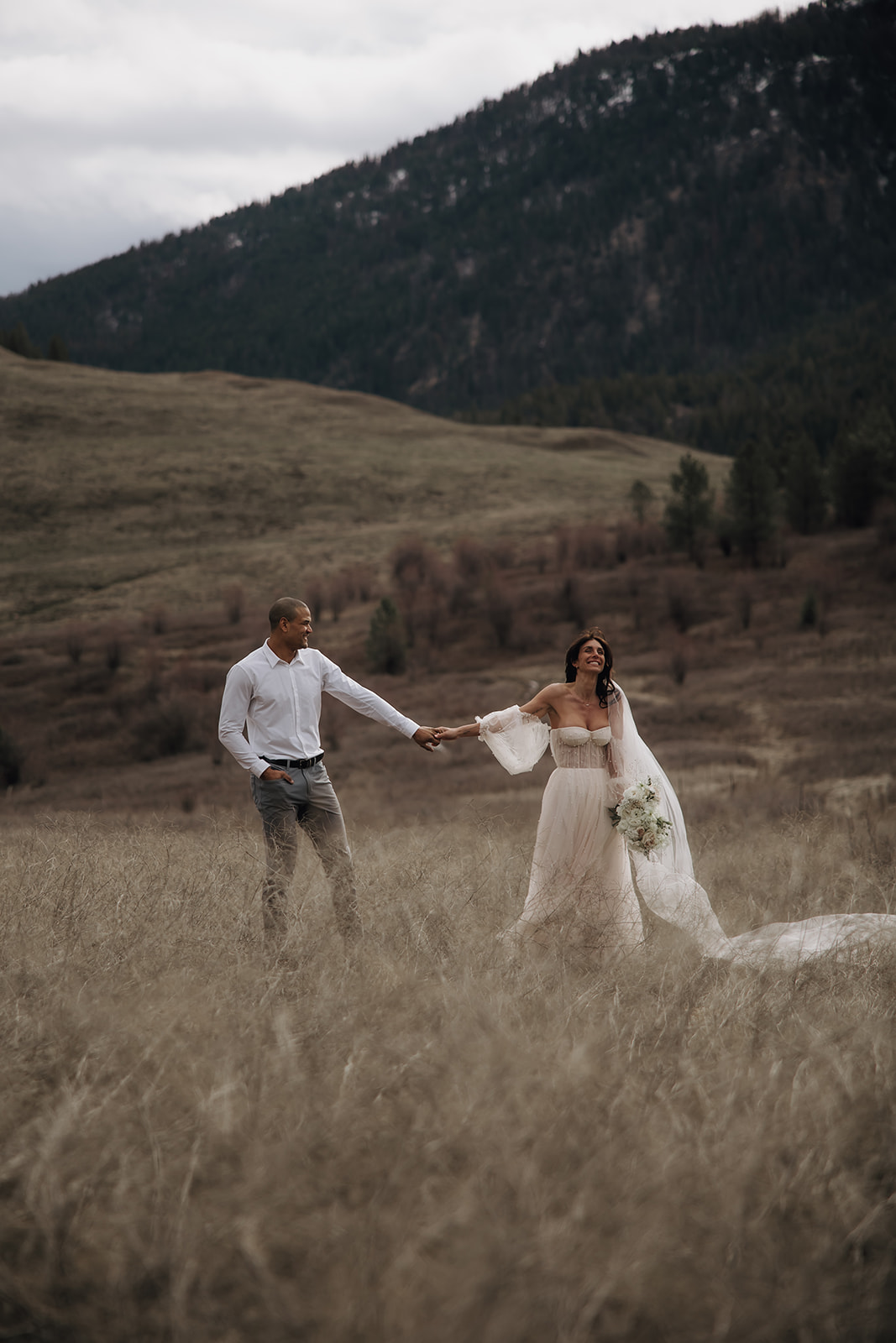 Couple holding hands during their Okanagan elopement, walking through a grassy meadow with a mountainous landscape behind them. The bride’s veil blows in the wind.

