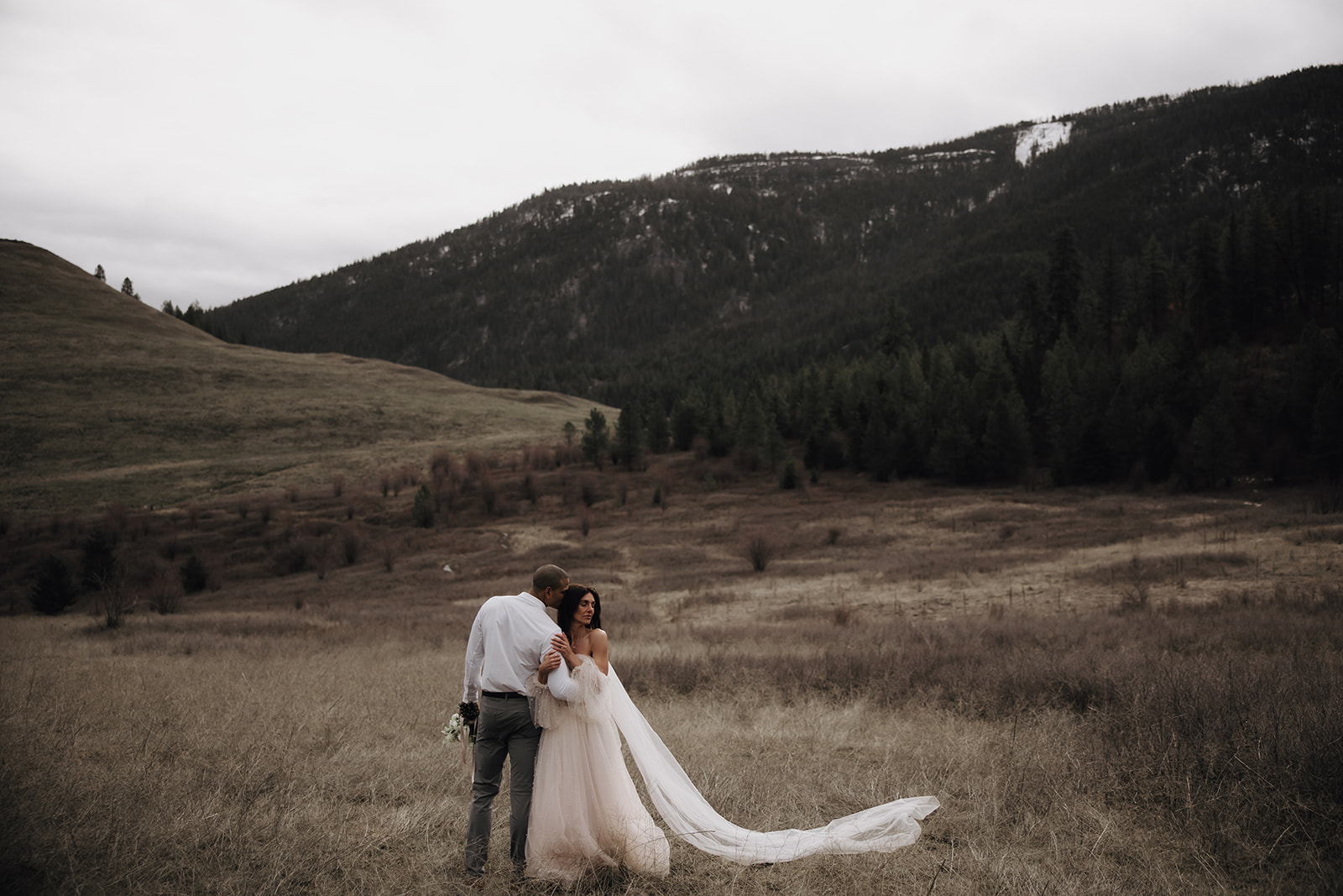 Couple embracing during their Okanagan elopement, surrounded by nature, with a mountain backdrop in the distance. The bride's veil flows gently in the breeze.