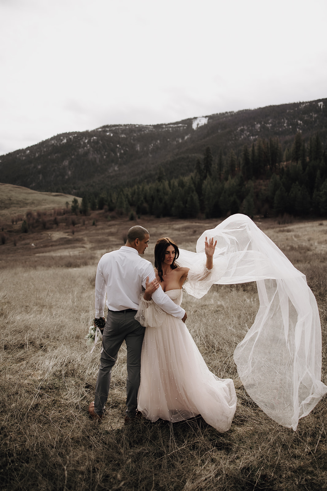 Couple holding hands during their Okanagan elopement, walking through a grassy meadow with a mountainous landscape behind them. The bride’s veil blows in the wind.

