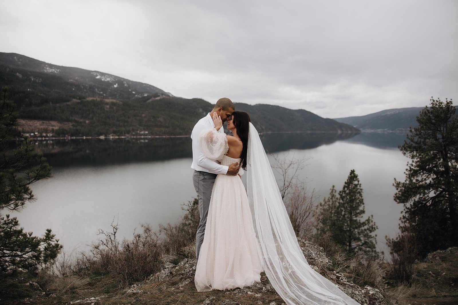 A couple embraces on a mountain edge, the bride’s long veil flowing in the wind, surrounded by the stunning landscape of the Okanagan during their Okanagan elopement.
