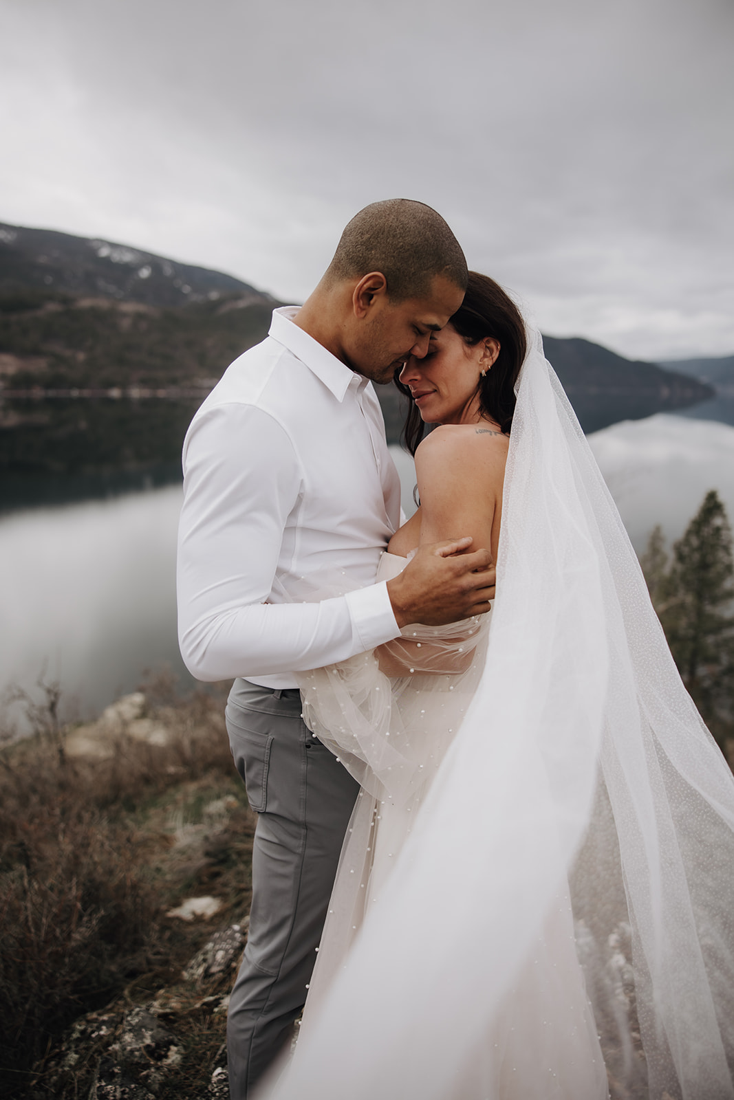 Couple sharing an intimate moment during their Okanagan elopement, the bride's veil flowing behind her as they stand against the backdrop of the mountains and lake.

