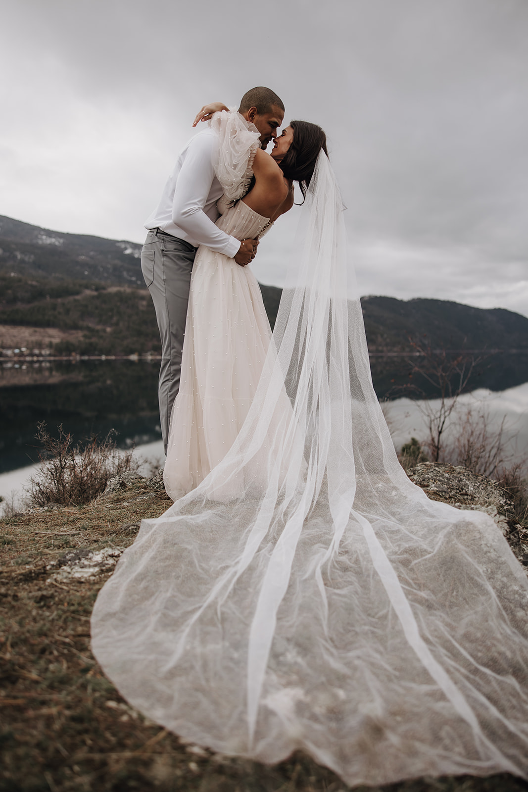 Bride and groom dancing together on a rocky outcrop, showcasing the beauty of their Okanagan elopement with a serene lake and mountain backdrop.

