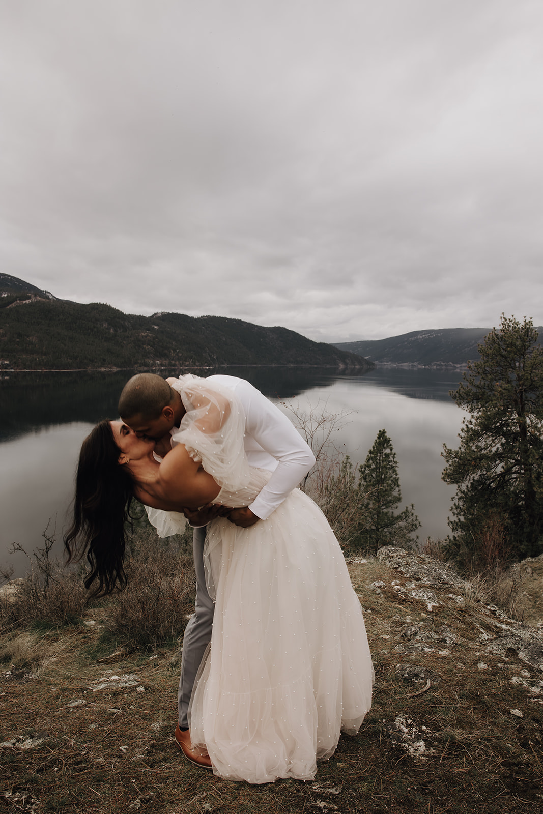 Couple embracing during their Okanagan elopement, surrounded by nature, with a mountain backdrop in the distance. The bride's veil flows gently in the breeze.

