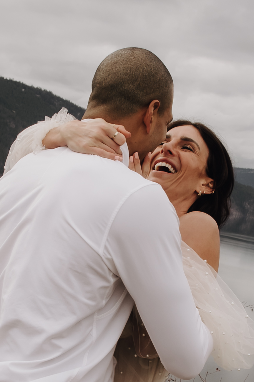 A couple shares a fun moment laughing together in a playful pose during their Okanagan elopement, with the lake and mountains providing a stunning backdrop.