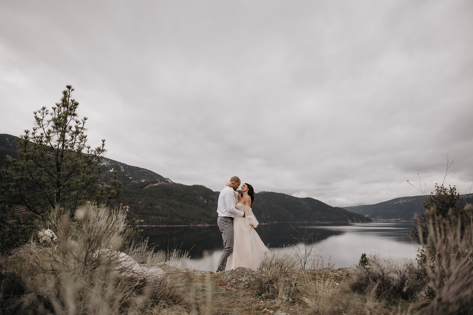 Couple embracing during their Okanagan elopement, surrounded by nature, with a mountain backdrop in the distance. The bride's veil flows gently in the breeze.