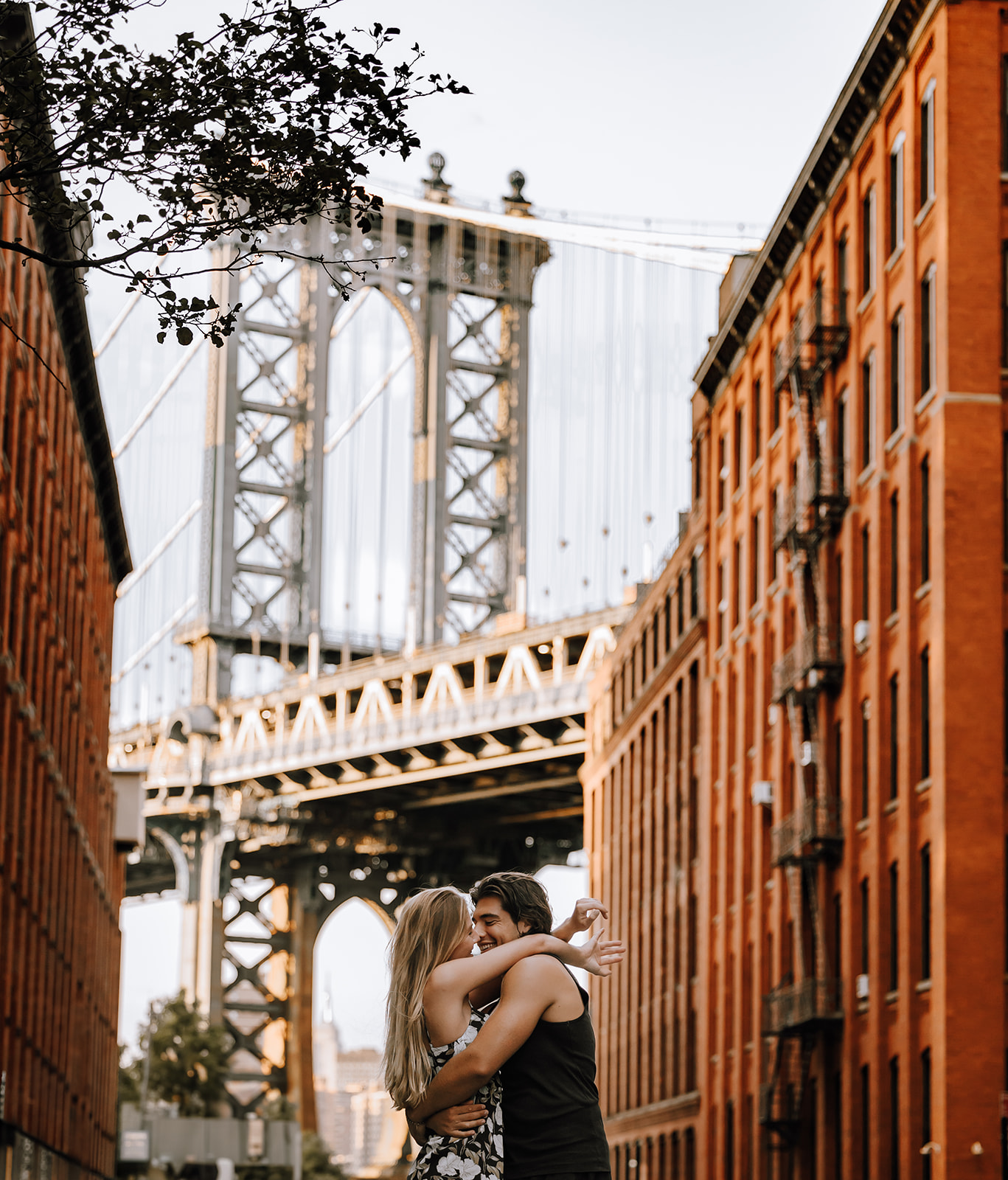 A couple embraces during their New York City couple photoshoot, with the Manhattan Bridge in the background and charming brick buildings framing the scene.