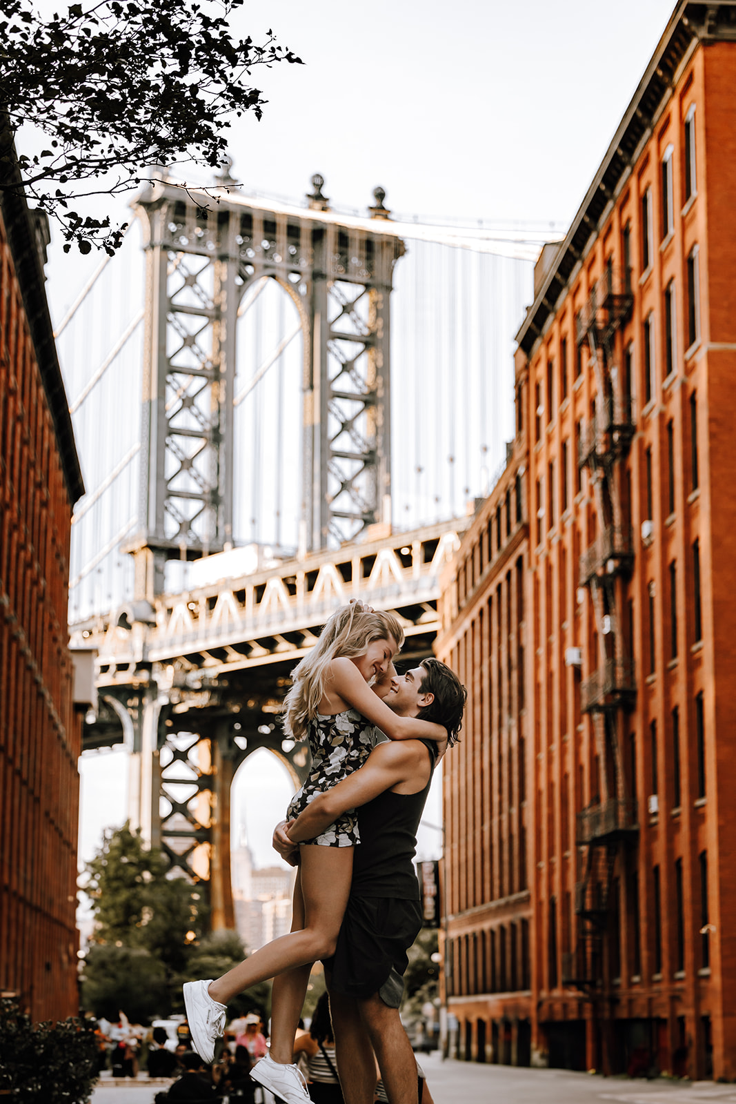 A joyful couple embracing in front of the Manhattan Bridge during their new york city couple photoshoot, with red-brick buildings in the background.