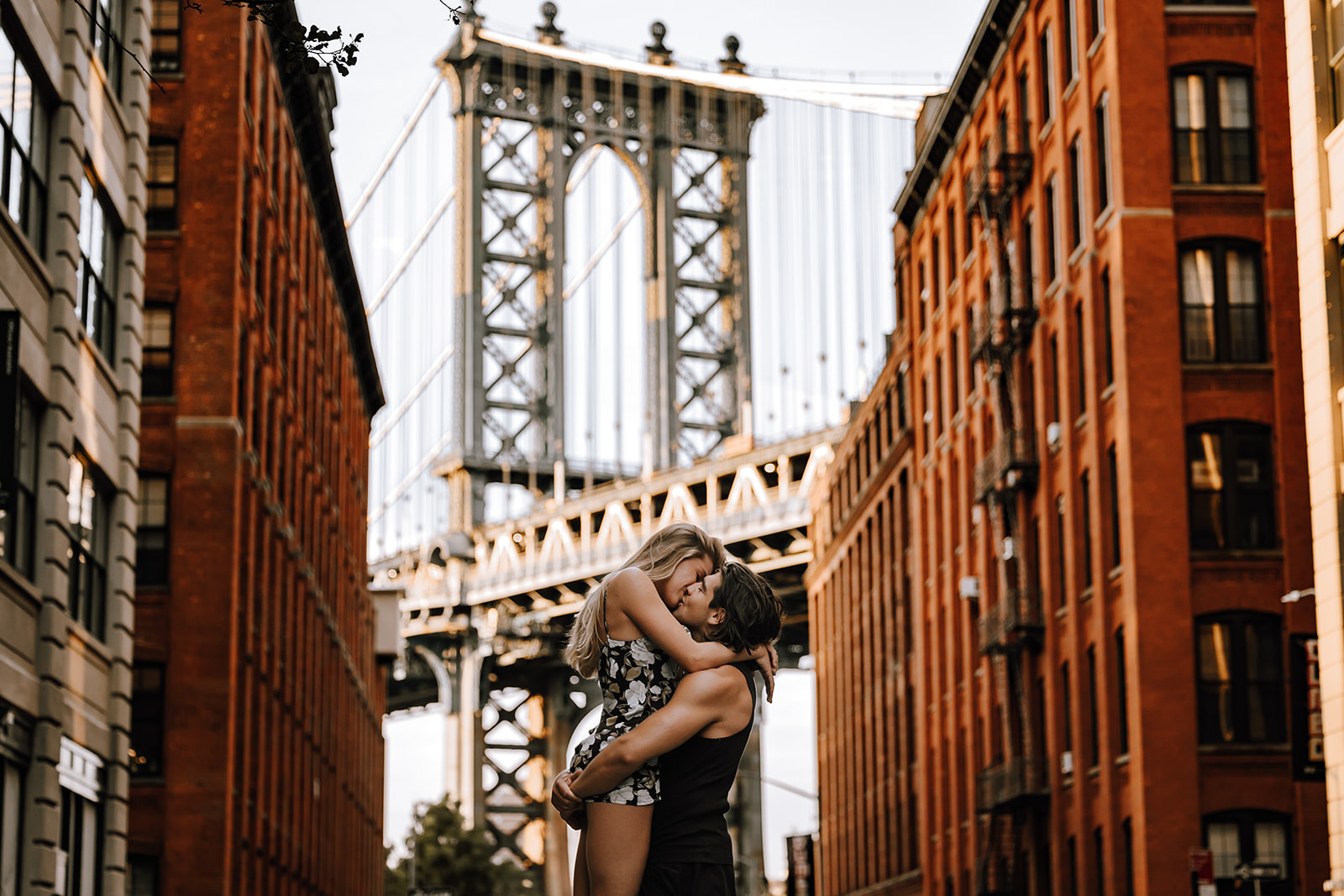 A joyful couple embracing in front of the Manhattan Bridge during their new york city couple photoshoot, with red-brick buildings in the background.