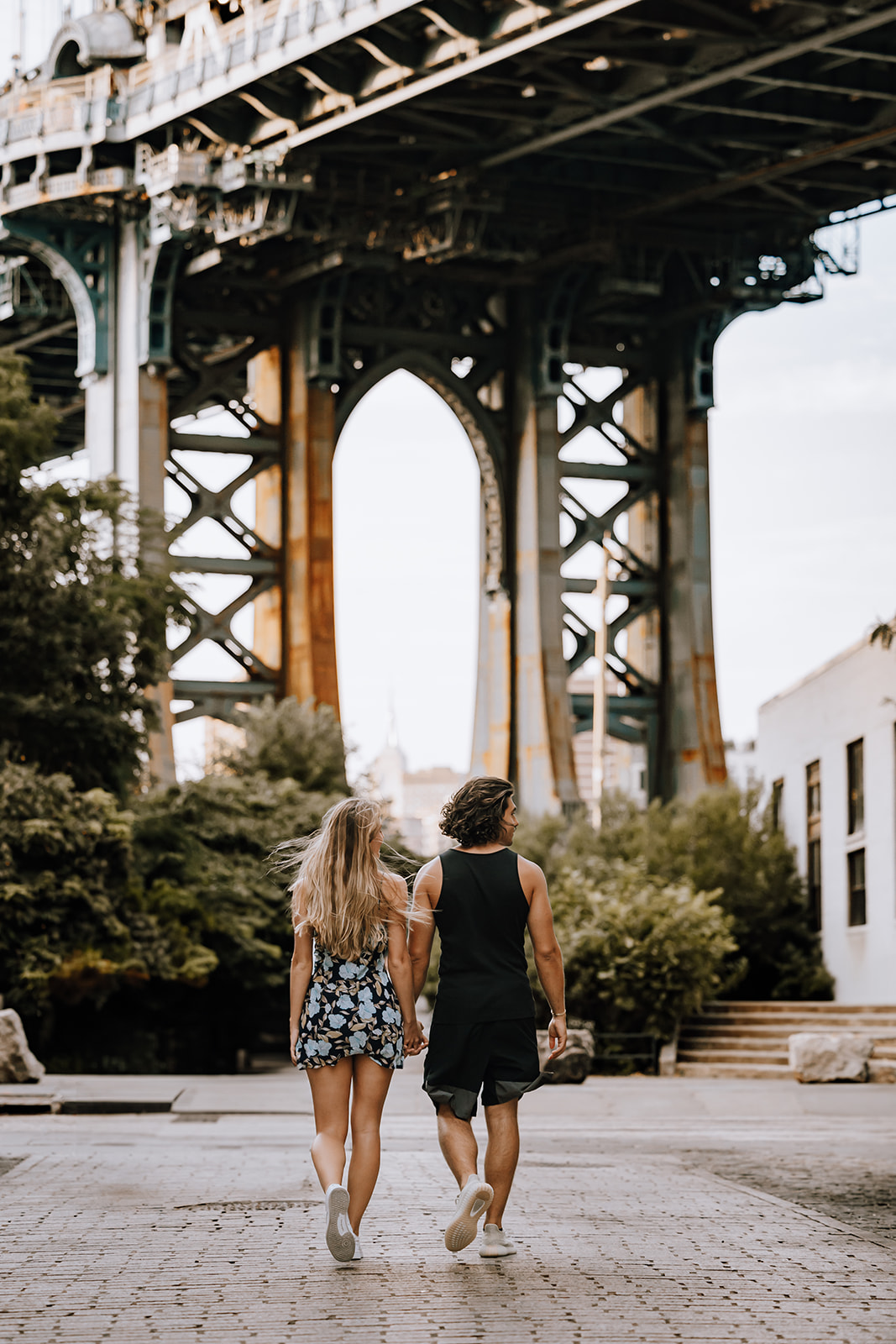 The couple walking away from the camera, hand in hand, under the majestic arch of the Manhattan Bridge during their city couple photoshoot.