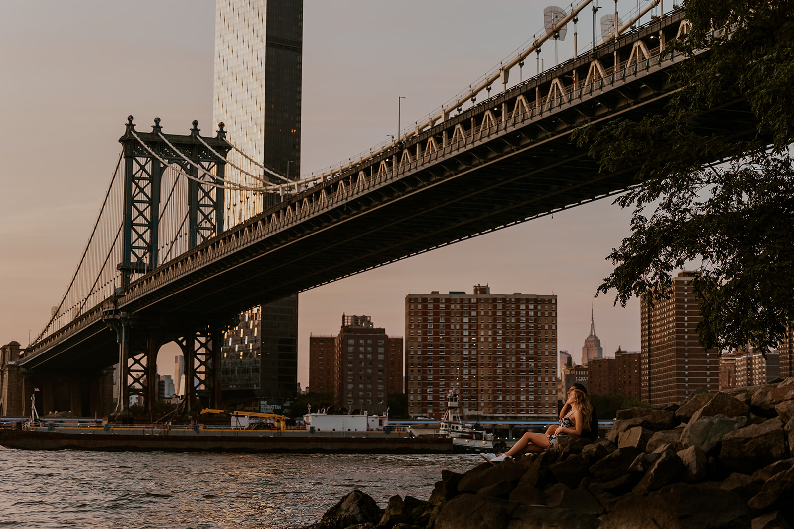 The couple sitting on a rocky shore and looking at the skyline during sunset, encapsulating the romance of their new york city couple photoshoot.