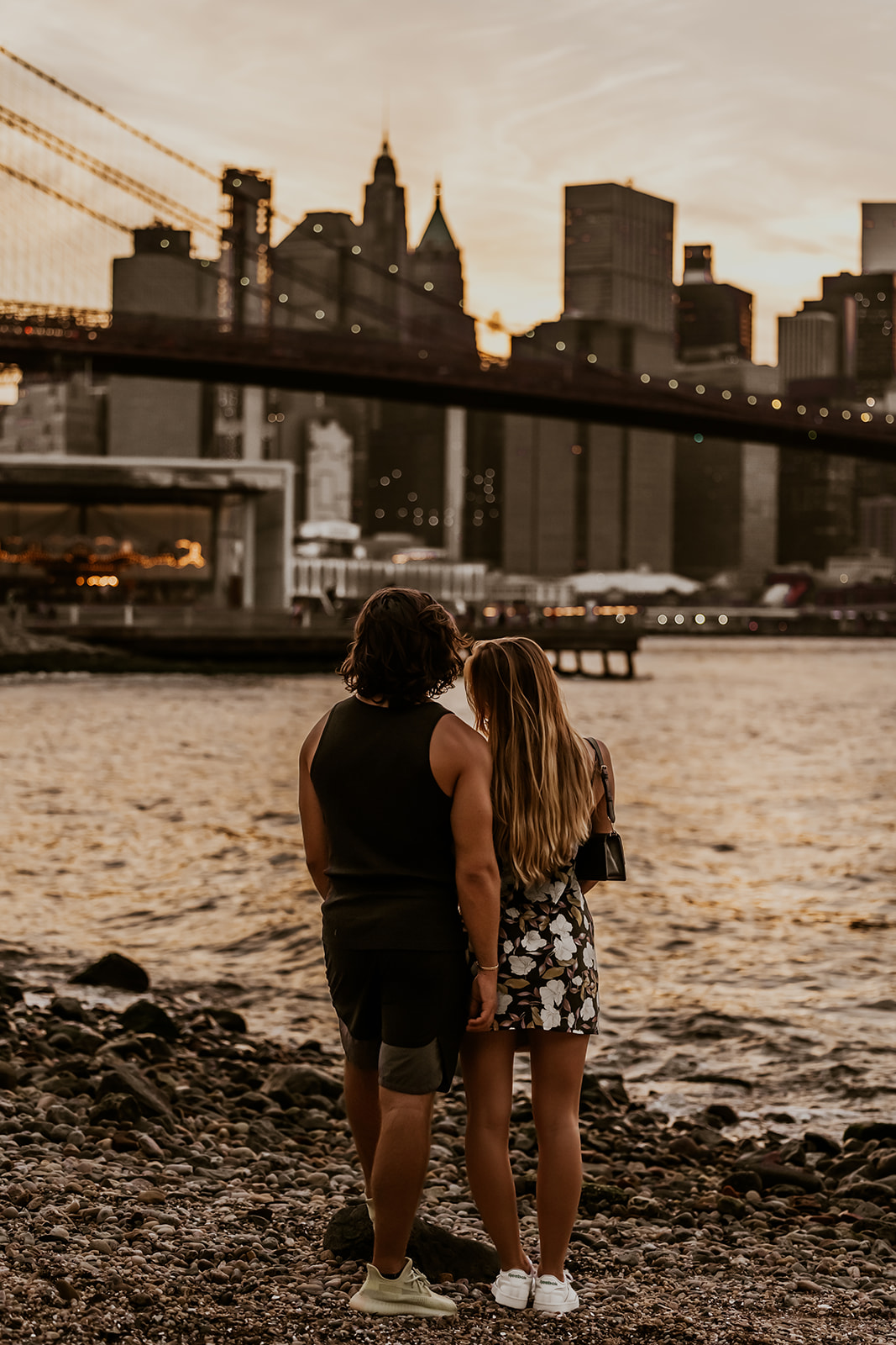 The couple standing on a rocky shore, holding hands and looking at the skyline during sunset, encapsulating the romance of their new york city couple photoshoot.