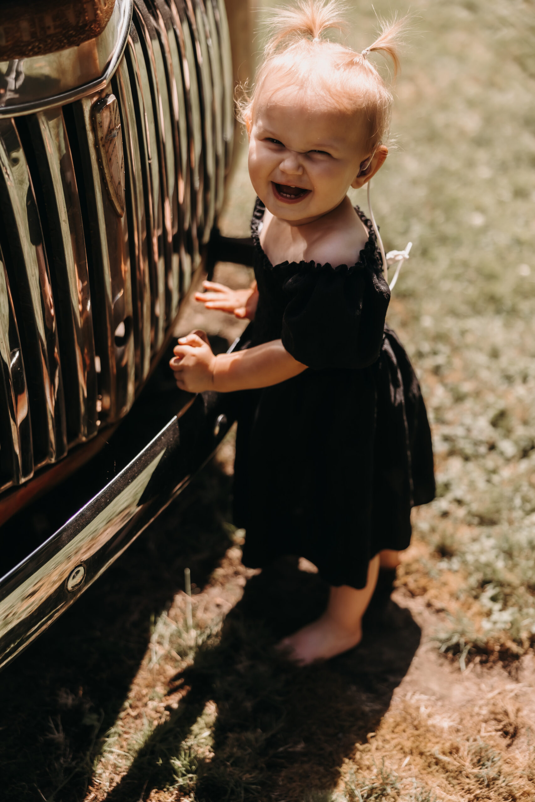 A little girl in a black dress grins as she plays by the vintage car at the wedding, her bright smile capturing the fun atmosphere of the Redwoods Golf Course wedding