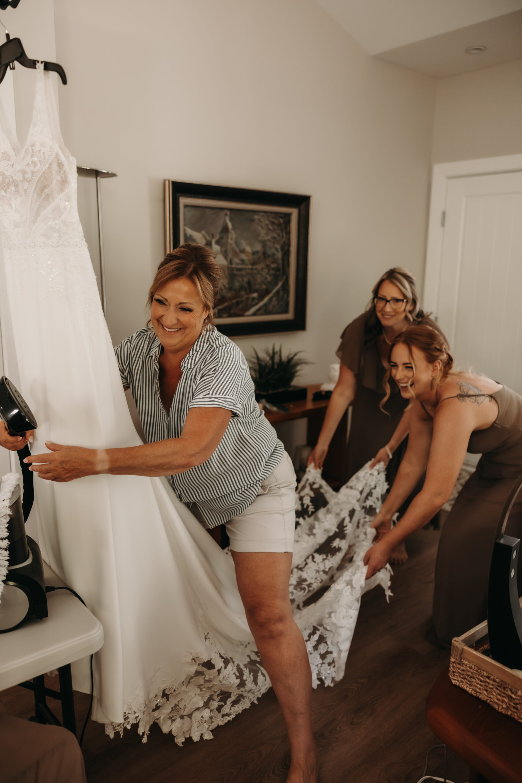 The bridal party prepares the wedding gown, with the bride's mother helping lift the dress. The attention to detail and happy energy of the moment shine through during this Redwoods Golf Course wedding.