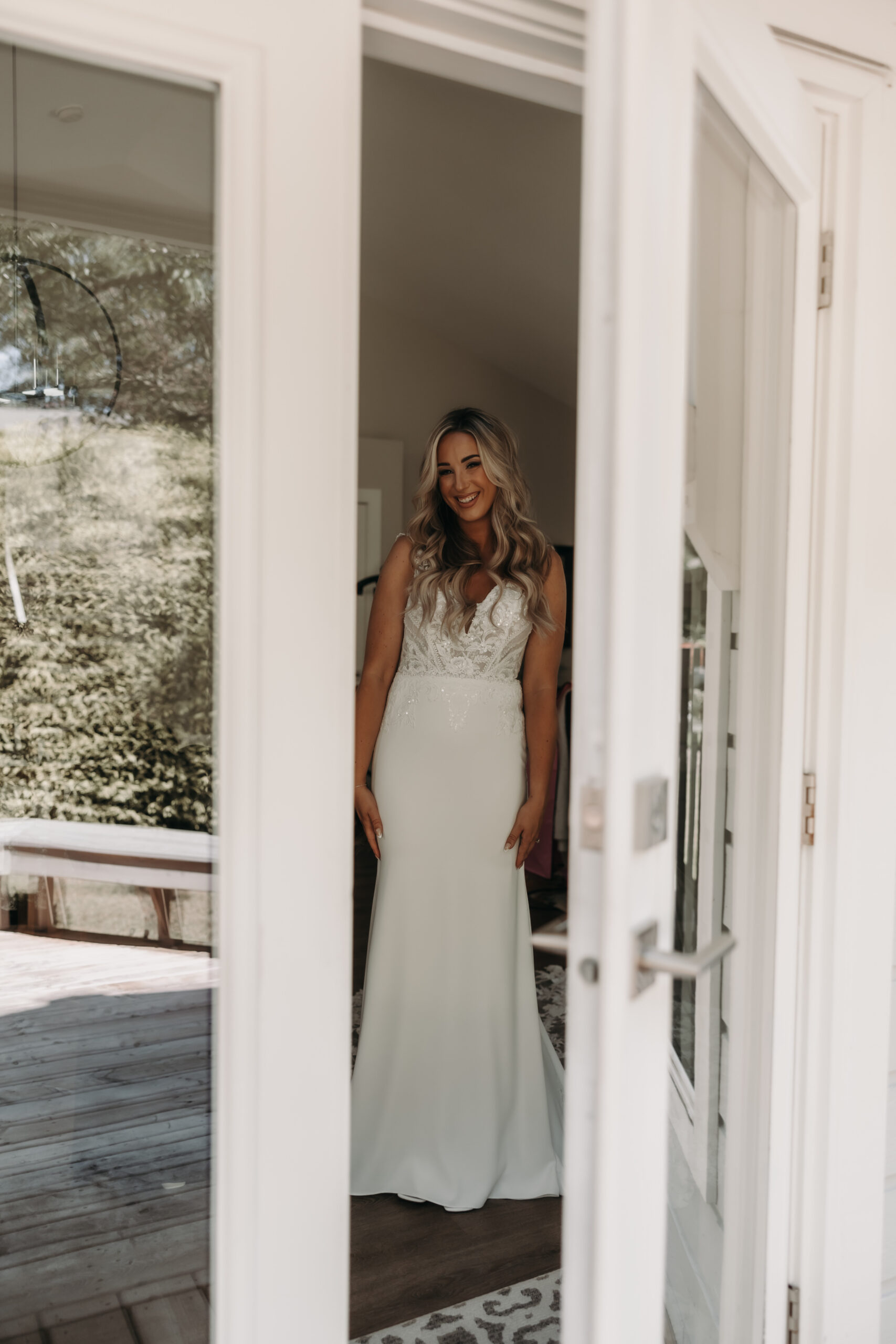 The bride stands in front of a door, smiling brightly as she looks at the camera. She is wearing her stunning wedding dress, with the reflection of the garden outside at the Redwoods Golf Course wedding visible in the glass door."