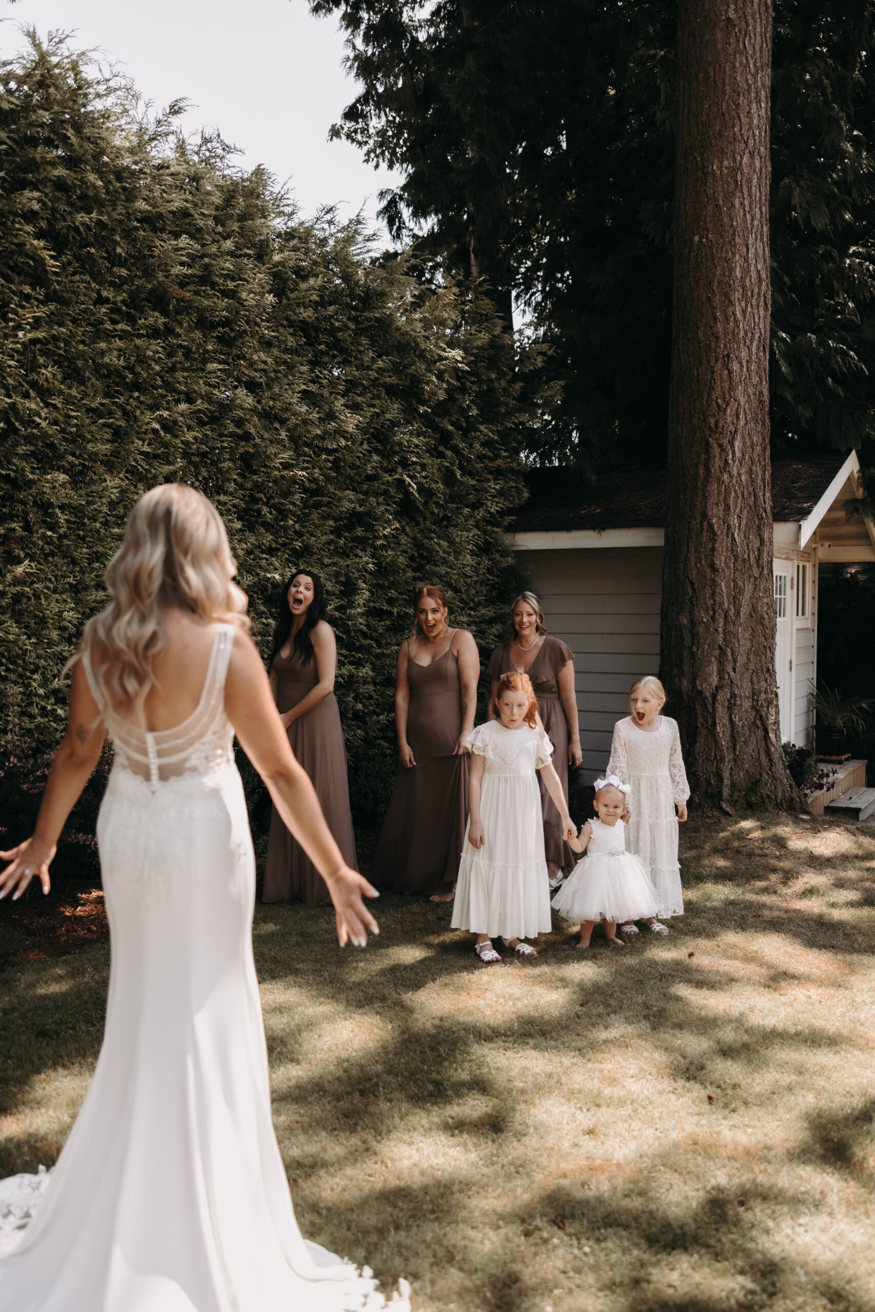 The bride walks toward a group of bridesmaids and children, all smiling and excited in their dresses. The lush greenery of the Redwoods Golf Course wedding venue sets the perfect tone