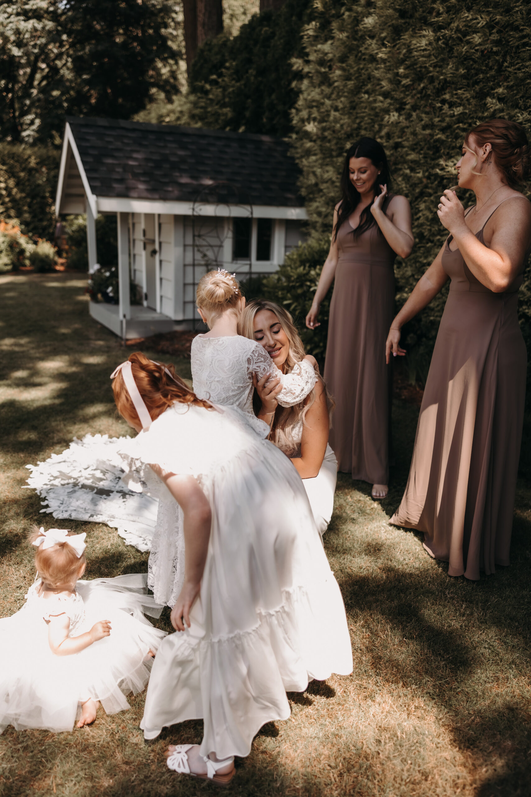 A bride hugs a child on the grass, with bridesmaids looking on in their brown dresses. The picturesque Redwoods Golf Course wedding backdrop adds a natural charm to the moment.