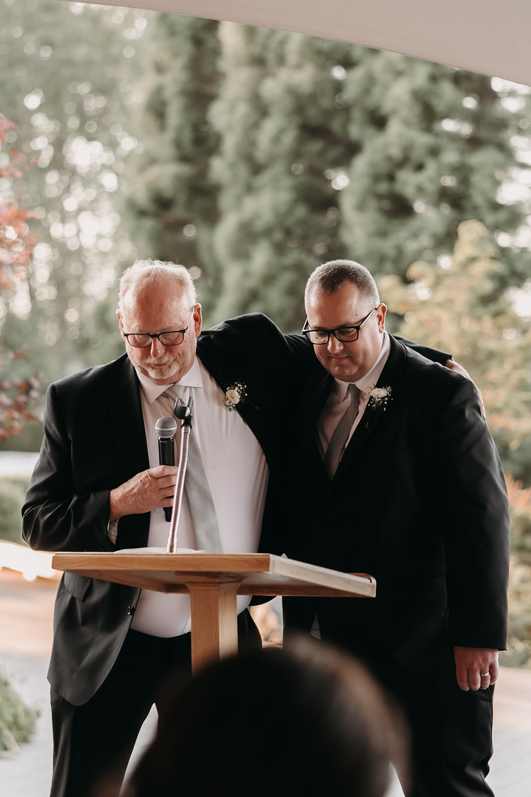 A father and adopted father stand together at the microphone, sharing a heartfelt moment during a wedding ceremony at Redwoods Golf Course Wedding, surrounded by lush greenery.