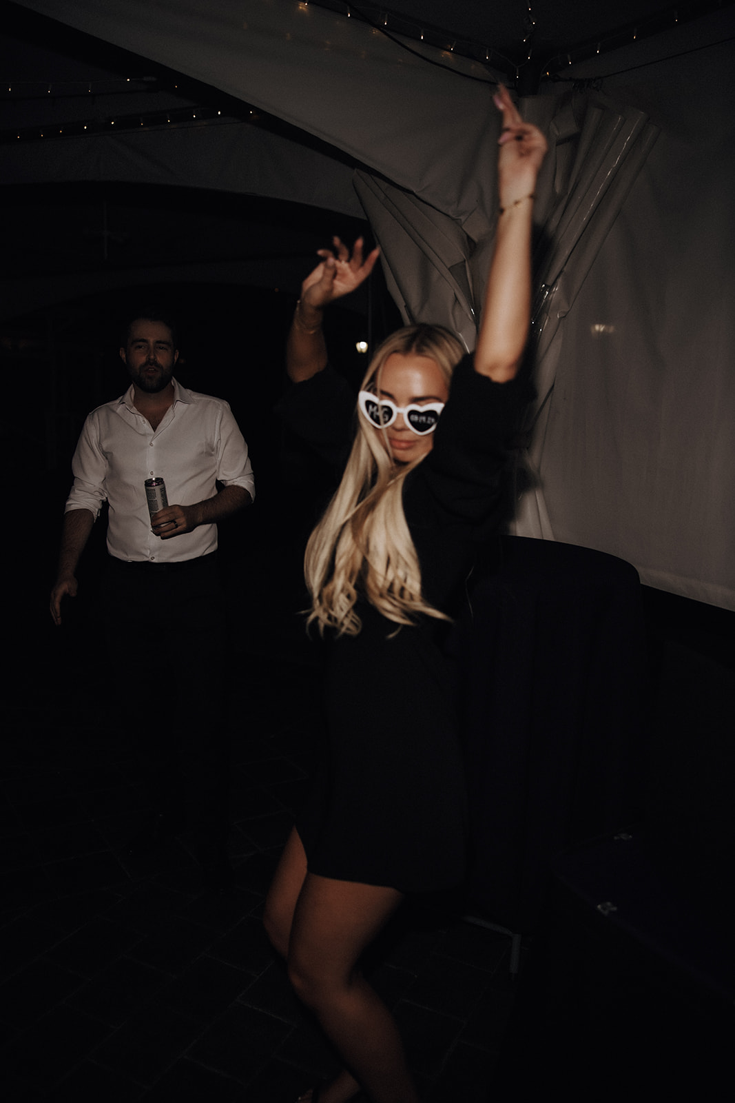 A guest dances joyfully at a wedding reception at Redwoods Golf Course Wedding, wearing fun heart-shaped glasses, with the groom's friend in the background.