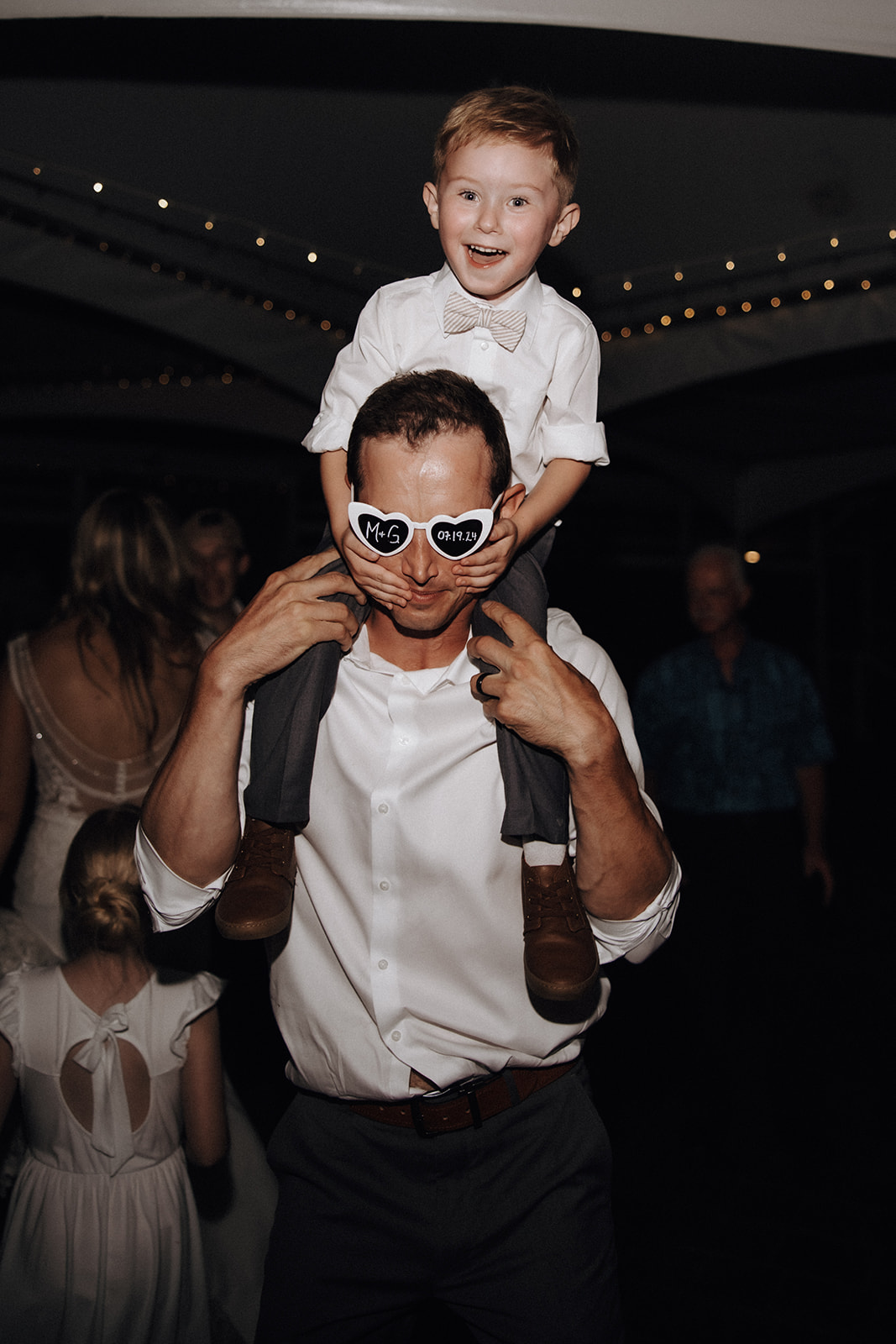 A playful and joyful photo of a little boy on his father's shoulders, both wearing heart-shaped "M+G" sunglasses, at the wedding reception at the Redwoods Golf Course.