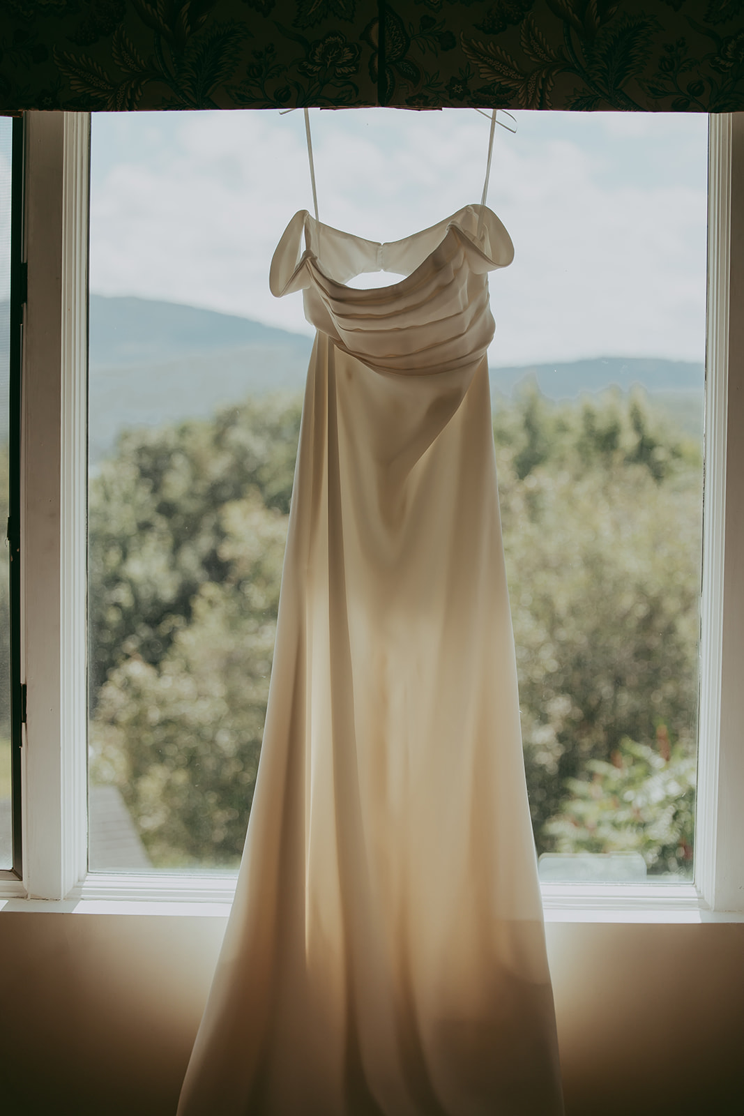 Bride’s dress elegantly hanging in a window, with breathtaking views of the Maine countryside, showcasing the beauty of a destination wedding in Maine