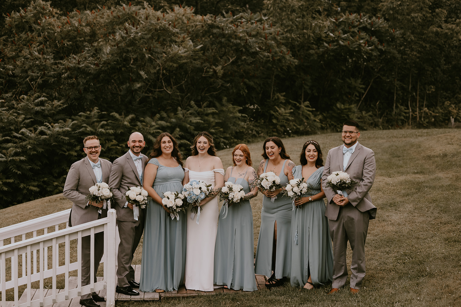 The bride and groom, surrounded by their wedding party, pose for a joyful group photo at their destination wedding in Maine, showcasing their stylish attire and beautiful bouquets.