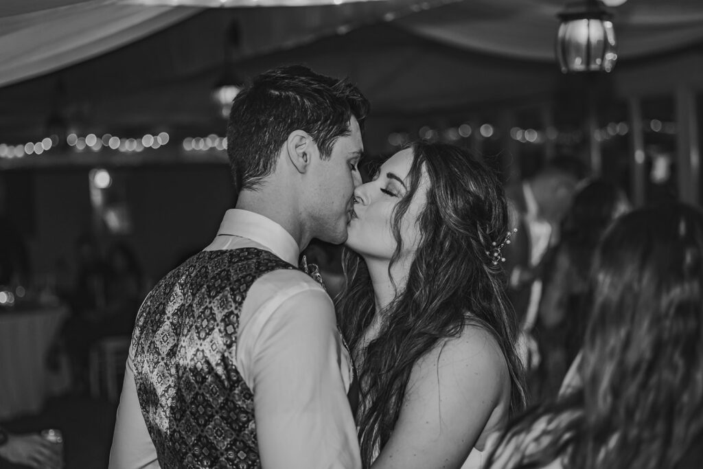 The newlyweds share a romantic kiss on the dance floor during their destination wedding in Maine, surrounded by family and friends, under the twinkling lights.