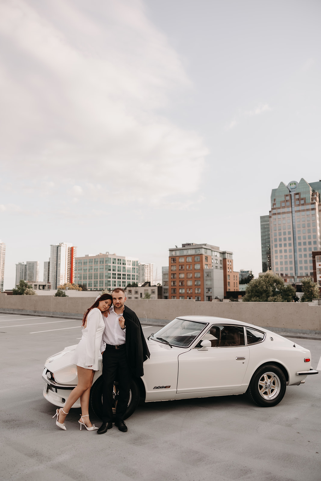 Couple posing together on a rooftop in Vancouver for city engagement photos, with a vintage car in the foreground and modern cityscape in the background.