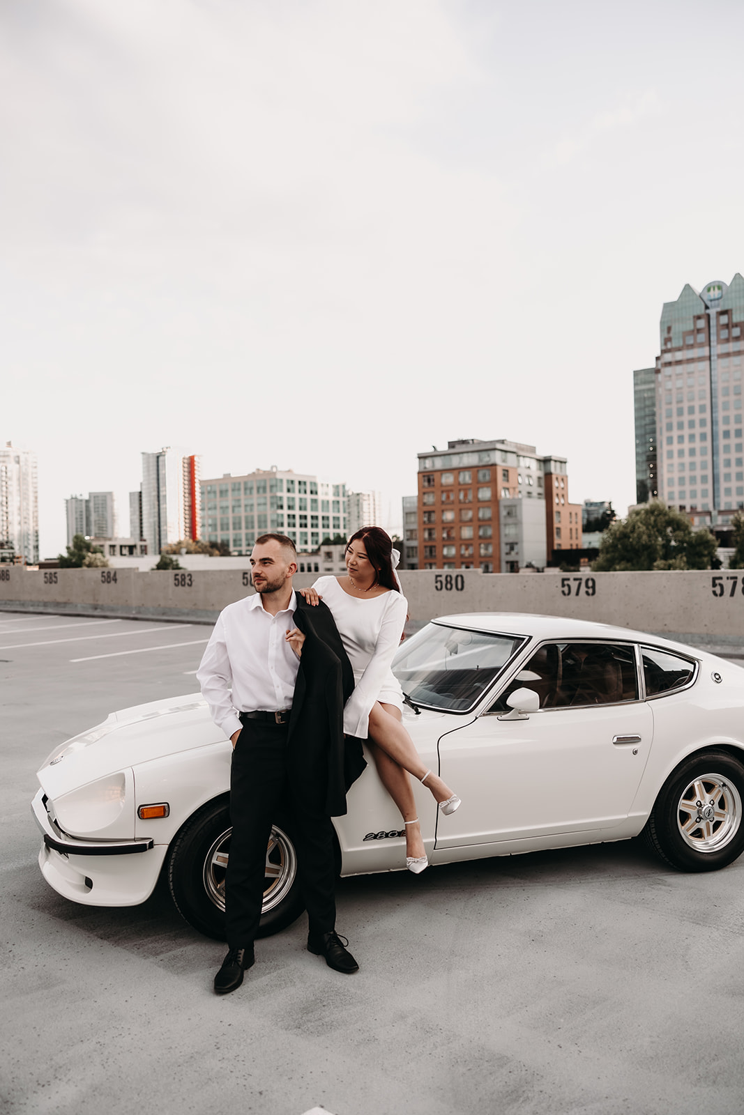 Engaged couple posing with a vintage car during their city engagement photoshoot in Downtown Vancouver, with modern city buildings as the backdrop.