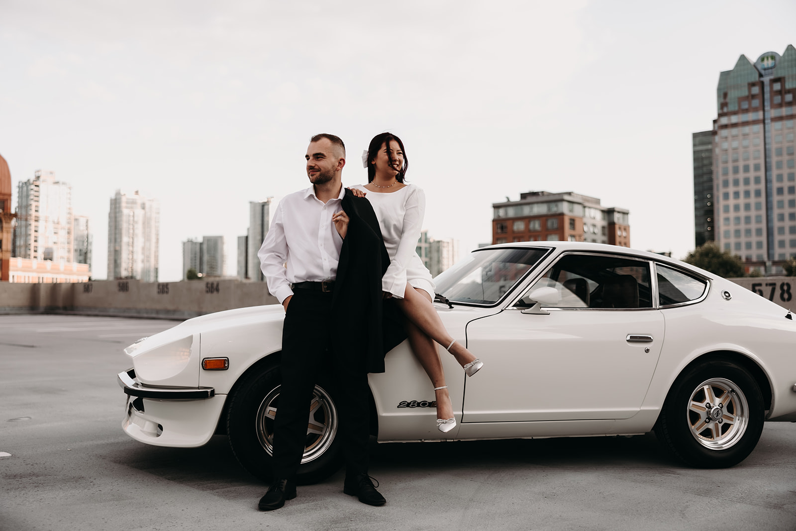 Couple laughing and enjoying their city engagement photos in Vancouver, standing close to a classic car on a rooftop with a beautiful sunset sky.