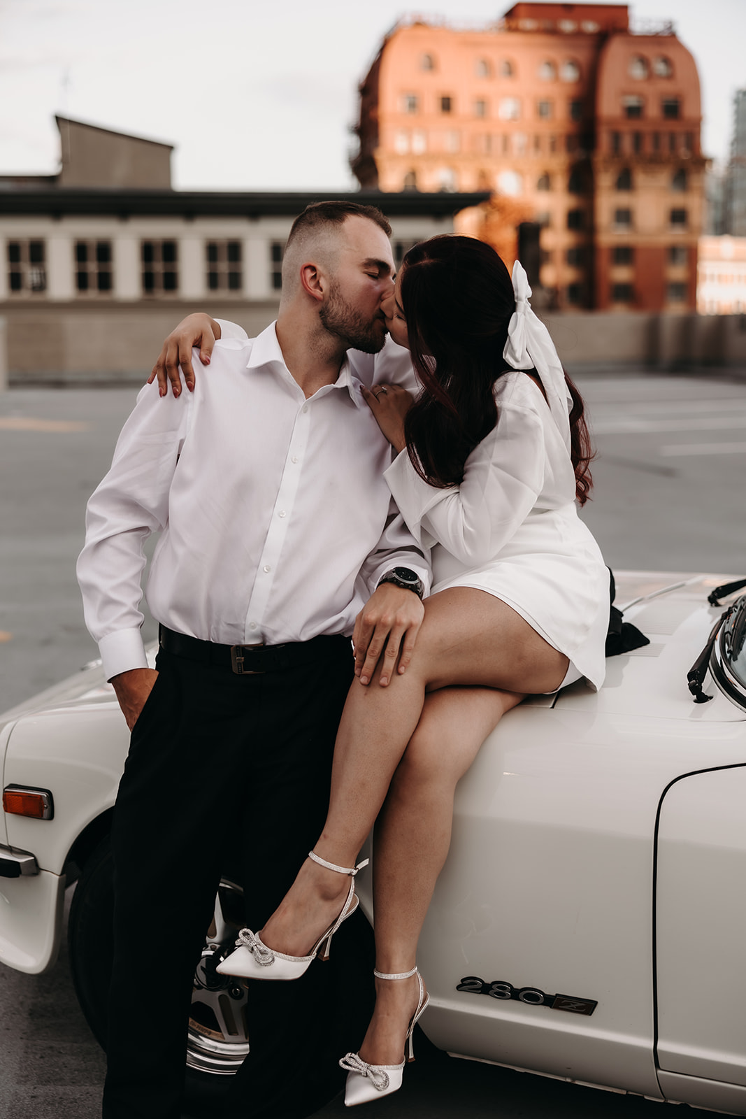 Couple sharing a fun, intimate moment during their city engagement photoshoot in Downtown Vancouver, with the vintage car and city buildings in the background.