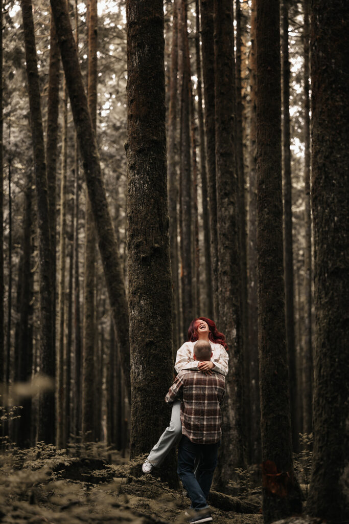 Engaged couple laughing as they embrace, surrounded by tall trees, showcasing their deep connection during their outdoor, forest engagement session.