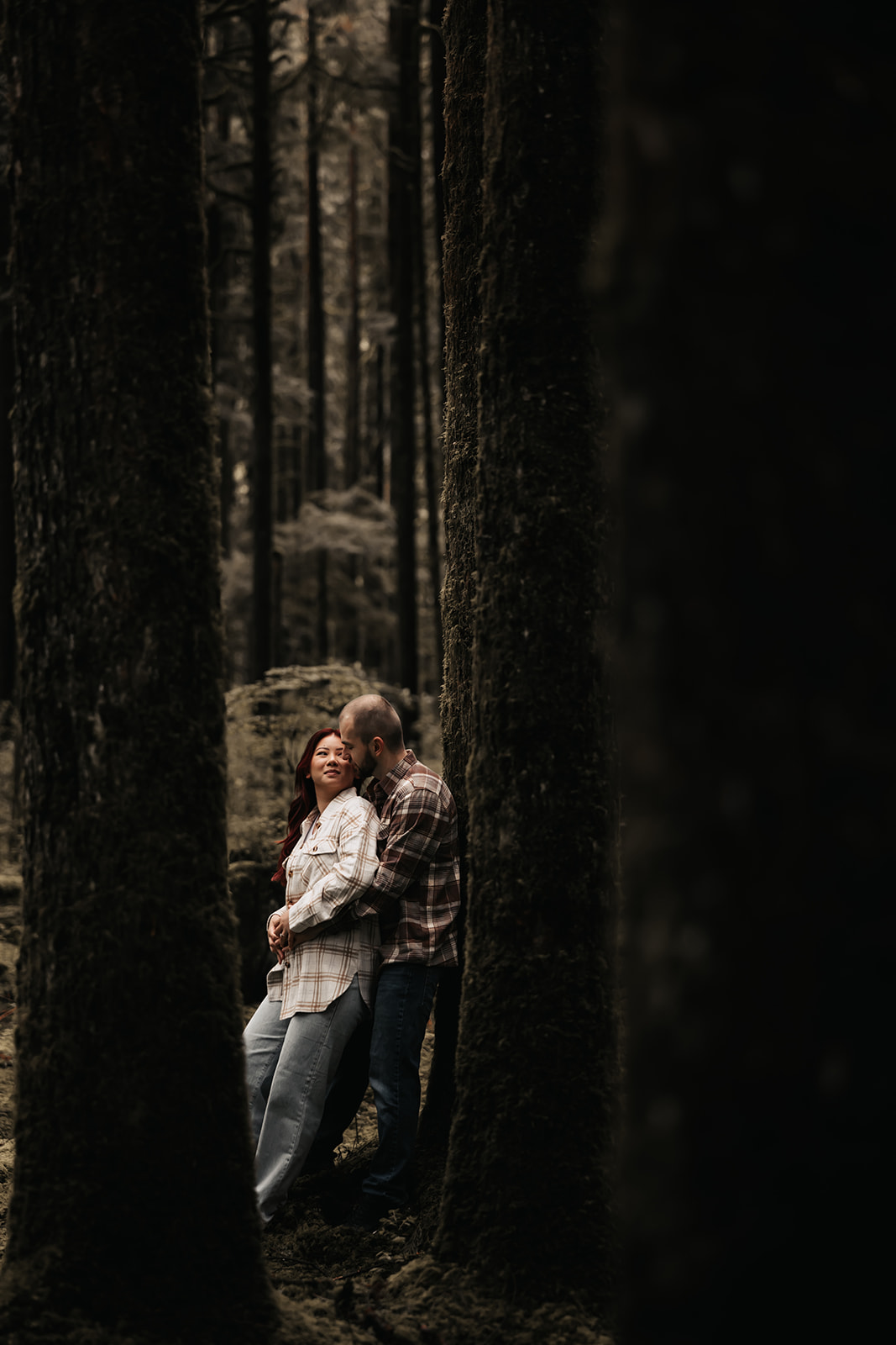 Romantic moment captured as the couple shares a sweet kiss in the middle of a lush forest with towering trees—a beautiful backdrop for rainy engagement photos.