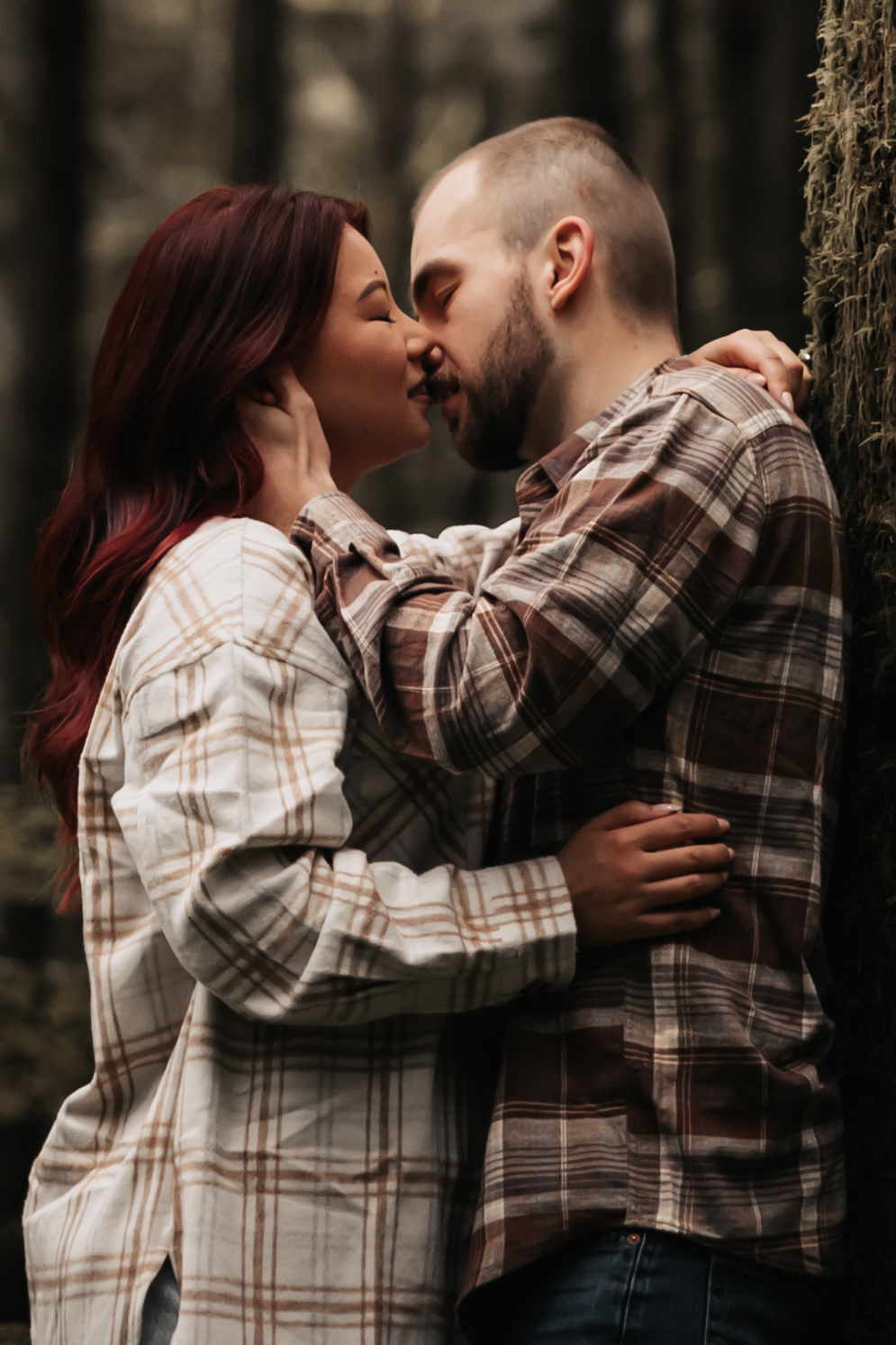 Engaged couple embracing in the forest, sharing a tender kiss surrounded by towering trees, a perfect example of capturing love in nature during rainy engagement photos