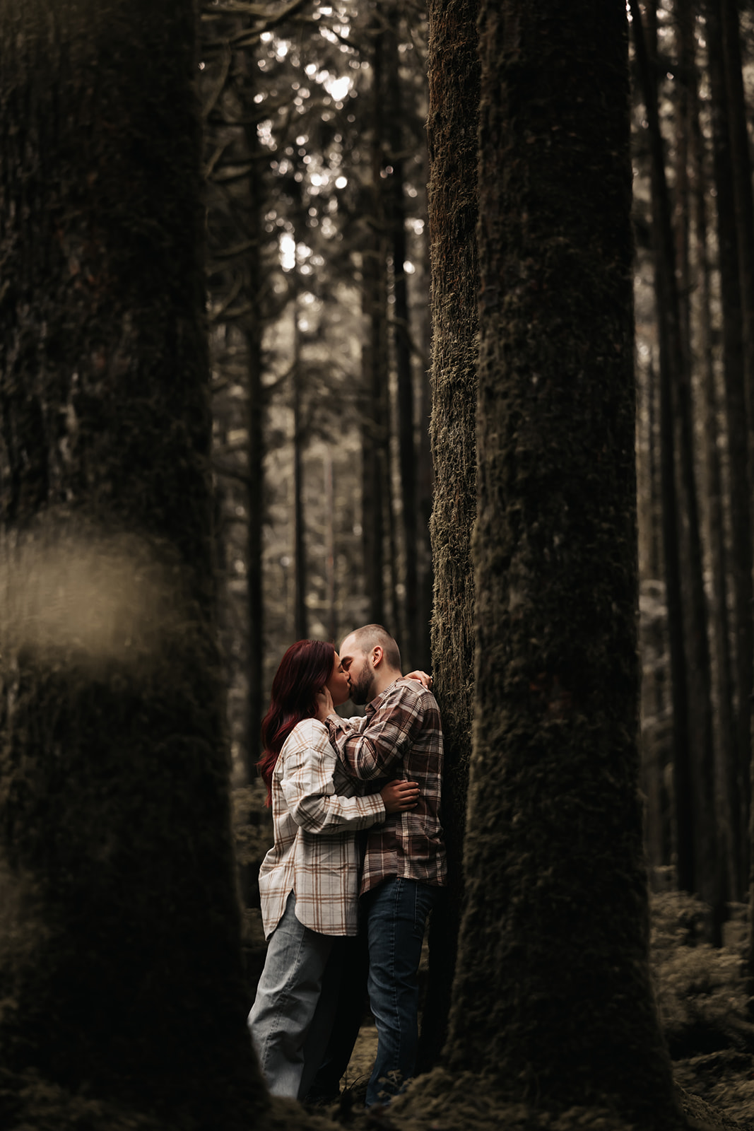 Engaged couple embracing in the forest, sharing a tender kiss surrounded by towering trees, a perfect example of capturing love in nature during rainy engagement photos