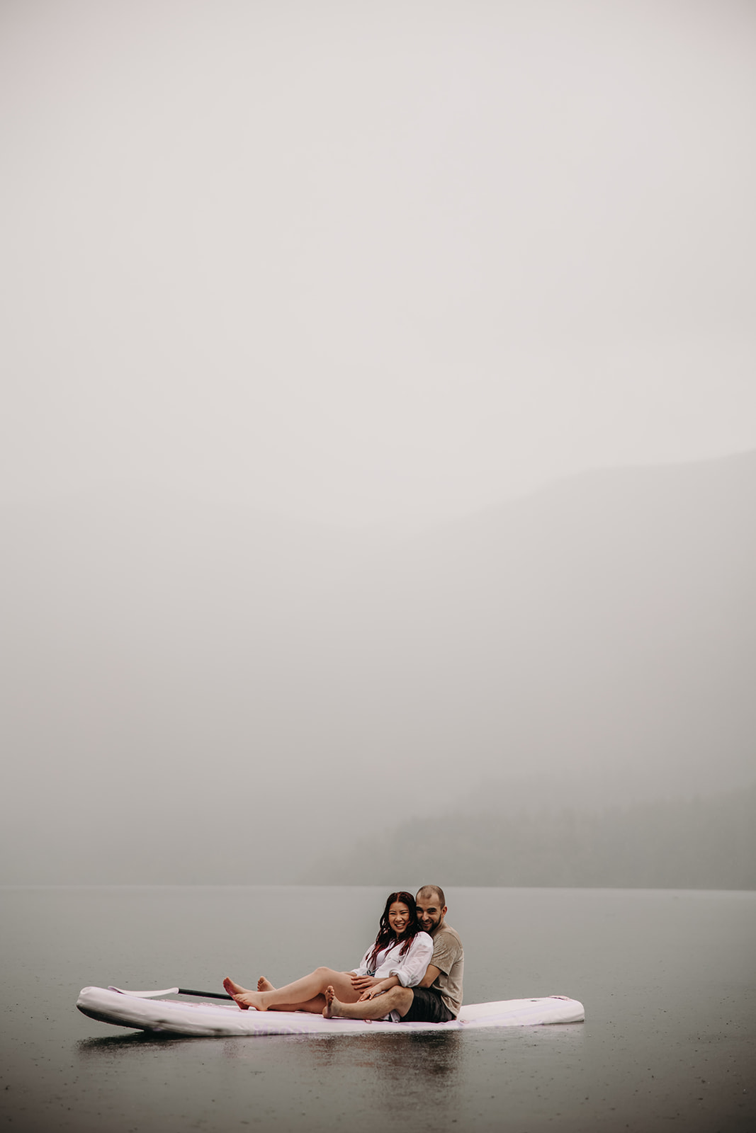 Couple sitting together on a paddle board during a rainy engagement photoshoot in BC.